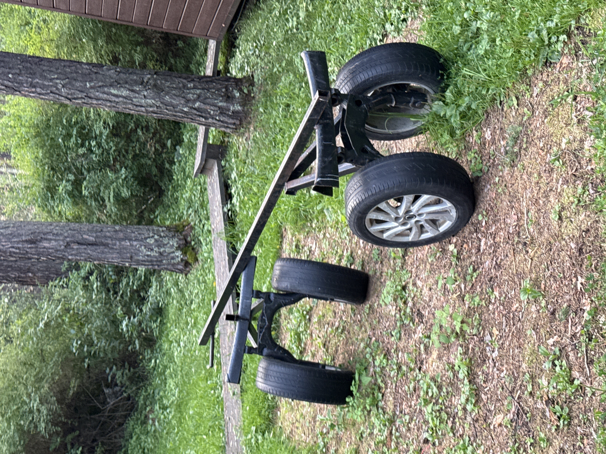 The image depicts a two-wheeled cart with a metal frame and four tires, situated outdoors on a dirt path with grass and scattered leaves. The cart appears to be stationary. Behind the cart, a wooden structure, possibly a building or deck, is partially visible on the right. Trees and dense green foliage form the background, suggesting a natural or semi-rural environment. The lighting indicates daylight. The location is identified as Barangol, Russia.