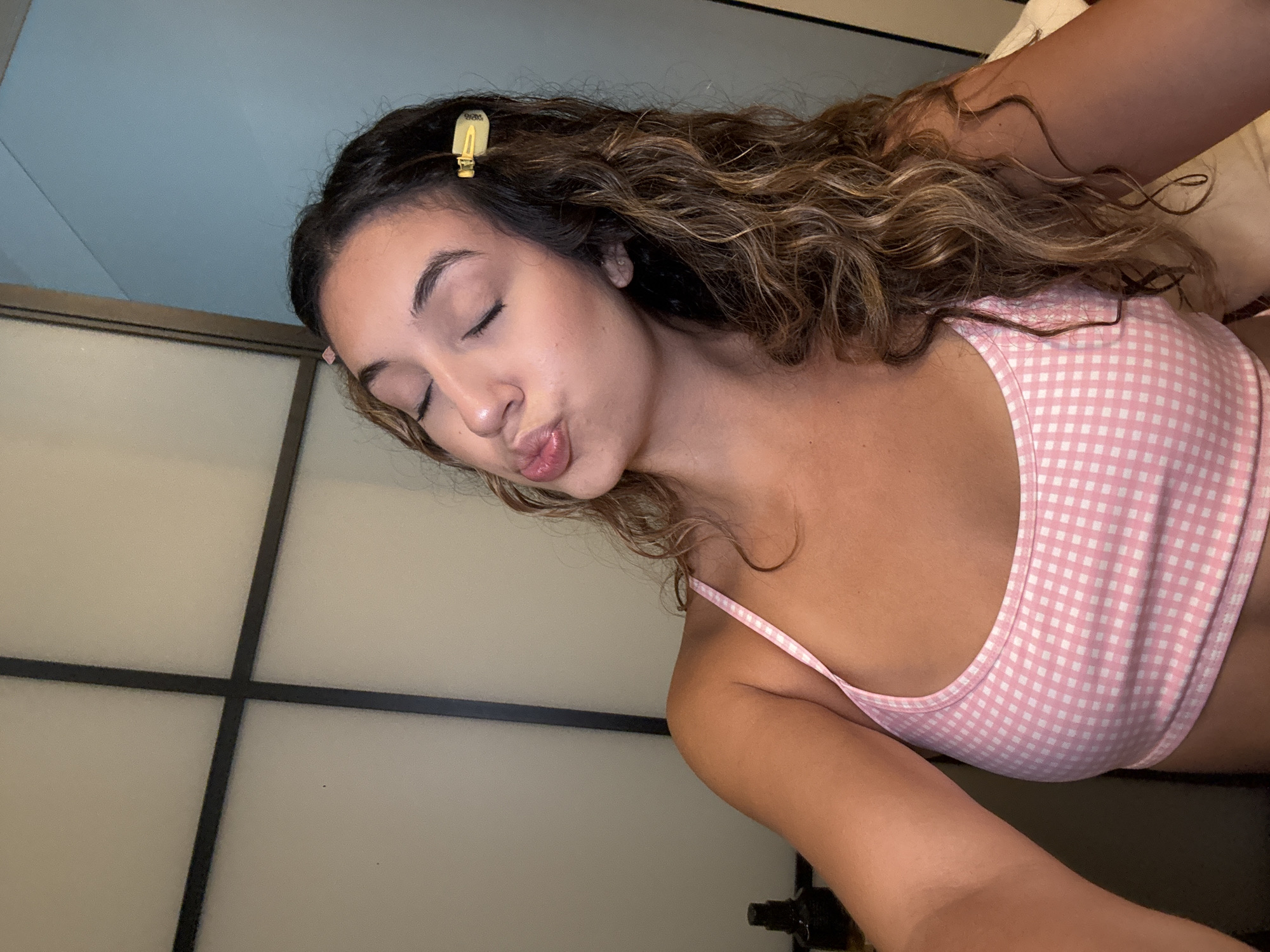 A woman with long, wavy brown hair and a pink gingham top is shown in a close-up shot. She has her eyes closed and her lips pursed in a kissing pose. A golden hair clip is visible in her hair. The background appears to be a wall with a grid design and a lighter colored section. The lighting is soft. The location is not specified.
