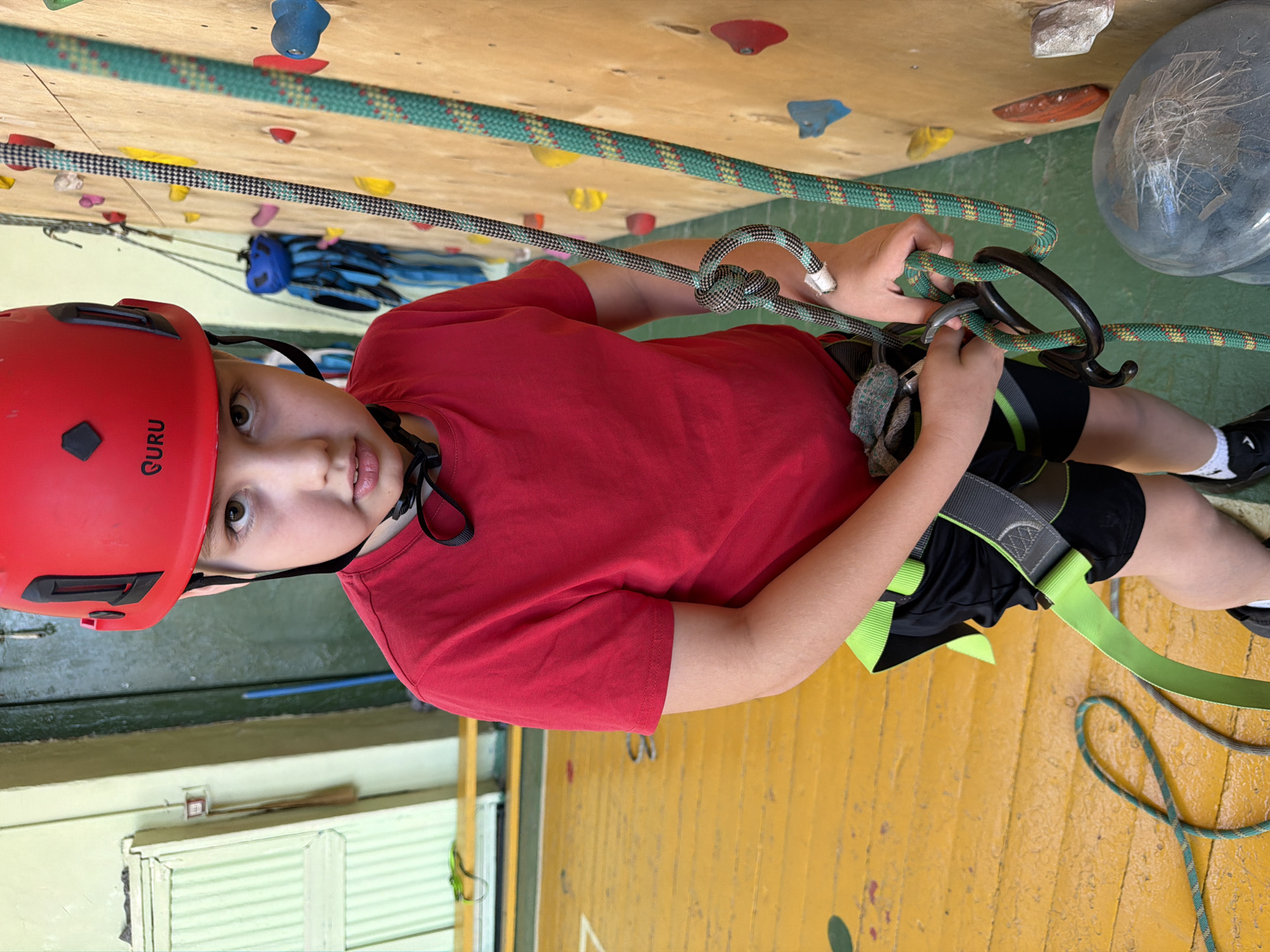 A young boy, wearing a red climbing helmet, a red shirt, and a climbing harness over black shorts, is indoors, likely in a climbing gym. He is positioned against a climbing wall with various colored holds, and two ropes are visible, one green and one black/gray. He is holding climbing equipment, seemingly attaching or detaching it. The wall beneath the climbing wall is yellow. To the left, there's a window with green and white slats.
It's impossible to verify the city of Izhevsk, Russia, without additional information.