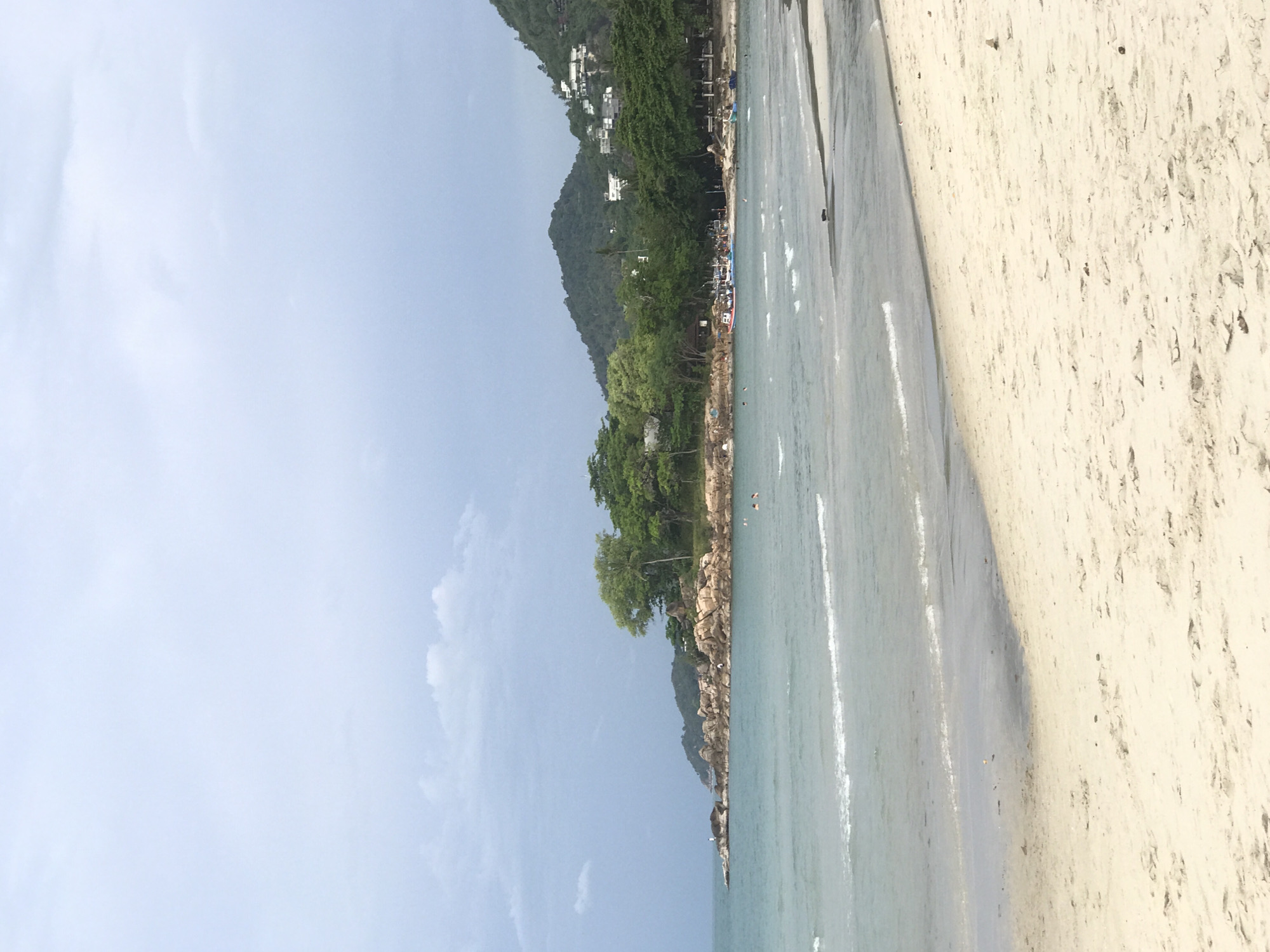 The image shows a beach scene in Baan Chaweng, Thailand. The sky is light blue with some clouds. Lush green vegetation covers a hill in the background, with a few buildings visible among the trees. The beach stretches from the right edge of the frame to the waterline. Clear, turquoise water meets the beach. Some people are visible on the beach and in the water.