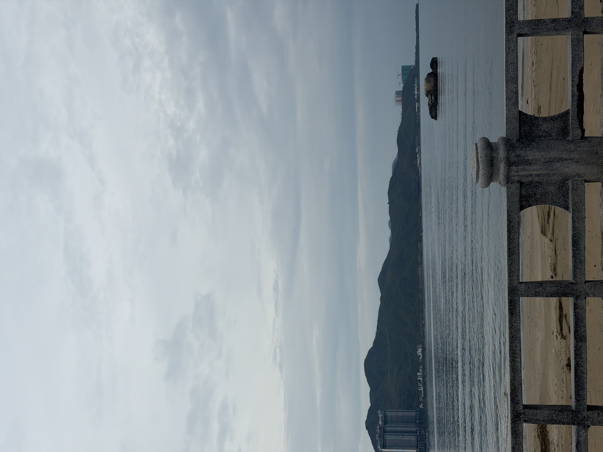 A landscape view of a sea bordered by mountainous terrain and buildings, framed by architectural details. The sky is overcast. The water is moderately choppy. A rock formation sits in the water, possibly an island or large outcropping. Buildings are visible near the shoreline at the base of the mountains. The right side of the image shows part of a grey structure, possibly a fence or balcony. The scene evokes Bac Nha Trang Ward, Vietnam.