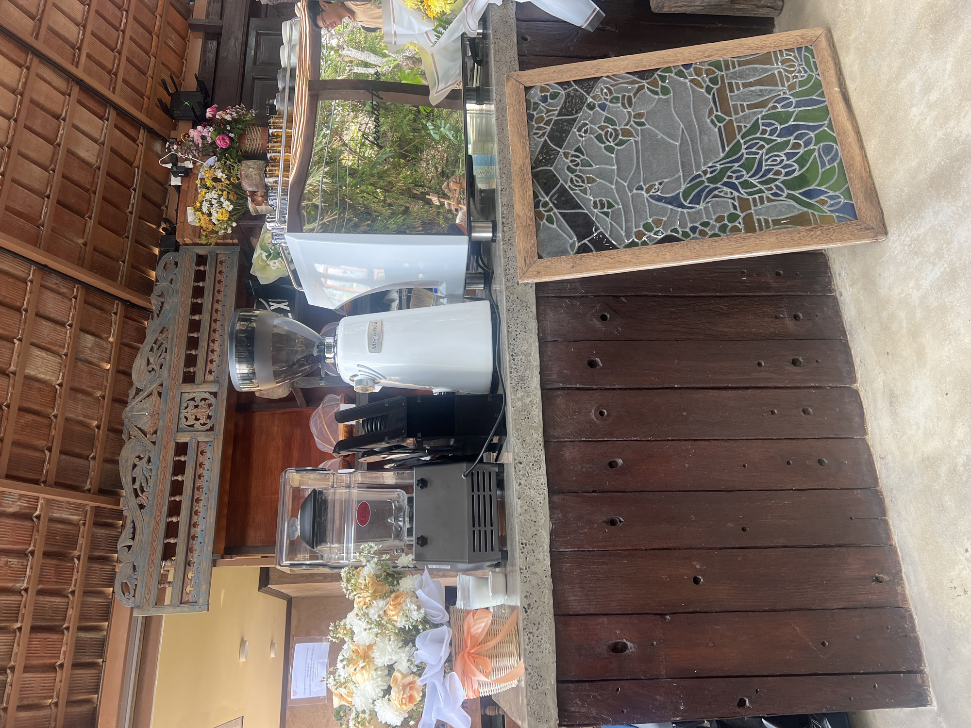An outdoor coffee bar counter with wooden planking and a concrete floor features a white commercial-grade coffee grinder and espresso machine. A blender sits to the left of the grinder. Behind the counter, floral arrangements are visible, along with a decorative carved wooden panel above. A framed stained-glass panel depicting a peacock rests on the floor in front of the counter. A person is visible in the background to the right. The scene appears to be in Kediri, Indonesia.