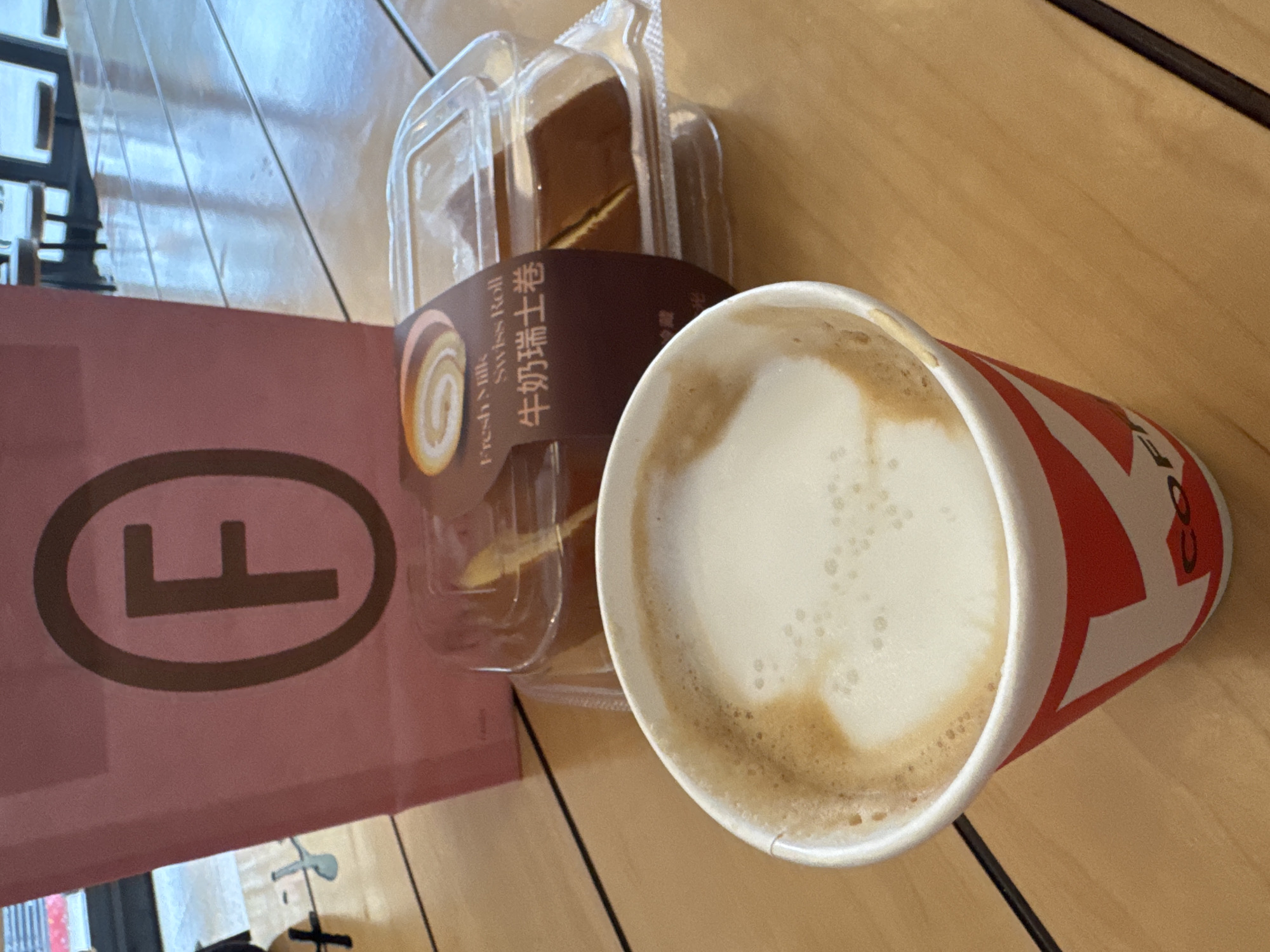 The image depicts a table with a coffee cup, a pink bag with a logo, and a packaged swiss roll. The packaging of the swiss roll is labeled in Chinese. The coffee cup is red and white, suggesting it is from a recognizable chain. The table appears to be wooden. The context of Qianjiang, China, is difficult to infer from the image alone without any landmarks, signage, or other locale-specific details.