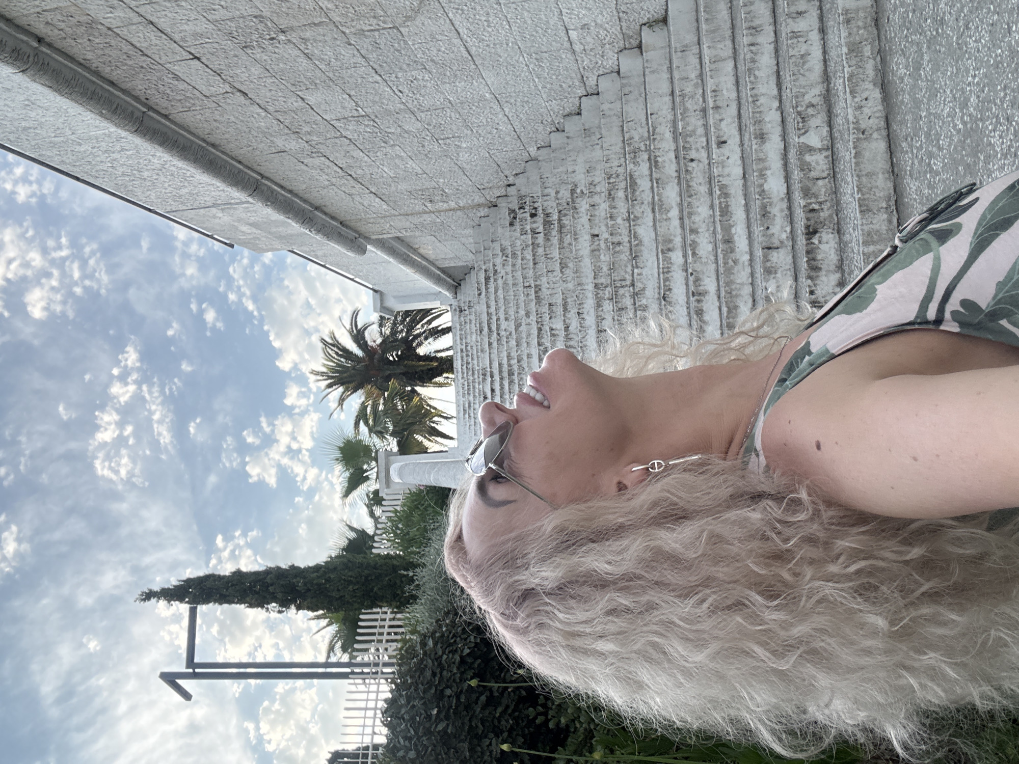 In Sochi, Russia, a woman with blonde, curly hair is captured in a low-angle, medium shot, looking up and to her right. She wears sunglasses and a patterned sleeveless top. The background features a large, white stone staircase with a textured railing ascending to the right. To the left, a tall, slender evergreen tree stands next to a white fence and lush green bushes. The sky above is partly cloudy with scattered white clouds against a blue backdrop.