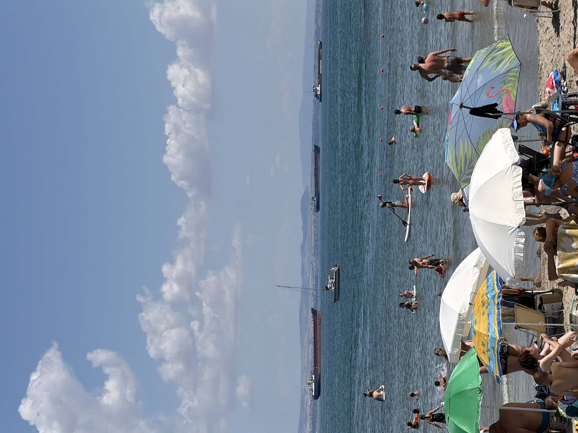 The image depicts a sunny day at a crowded beach in Burgas, Bulgaria. Several people are enjoying the water and sand. In the foreground, beach umbrellas, including one with a flamingo design, offer shade to sunbathers. People are seated on chairs and towels on the sand.
The sea is populated with numerous individuals engaged in various activities. Some are swimming, while others are paddleboarding. A few people are standing in the shallow water. In the background, large cargo ships are visible on the horizon, suggesting the location is near a shipping route or port. The sea is calm, and the sky is bright blue with scattered white clouds. The overall scene conveys a sense of leisure and a typical summer day at the beach.