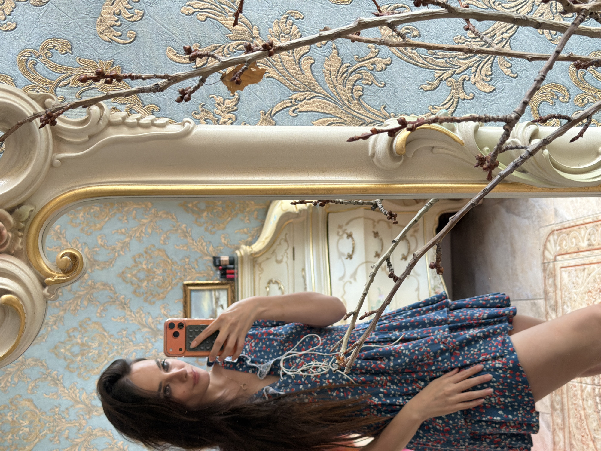 The media file is a vertical, eye-level, medium shot captured in a brightly lit room. The image is a reflection in a large, ornate mirror, which frames the scene. In the foreground of the reflection, a young woman is visible, taking a selfie with her orange-cased smartphone. She is wearing a dark blue, floral-patterned dress with short sleeves and a ruffled neckline. Her long, dark hair is down, and she is looking directly at the camera with a neutral expression. Her left hand is visible, resting on her dress.
The background of the reflection is a room with ornate, light-colored furniture and patterned wallpaper in shades of blue and gold. A framed picture hangs on the wall. In the actual scene, the mirror's frame is prominent, appearing as a large, cream-colored, carved structure with gold accents. Bare branches with small buds and a single dried leaf are draped across the top of the mirror, obscuring parts of the reflection and the wallpaper. The overall impression is one of an indoor, decorative setting. The lighting suggests daytime, and there are no clear indications of weather or specific time of day beyond that. No visible text is present in the media file.
