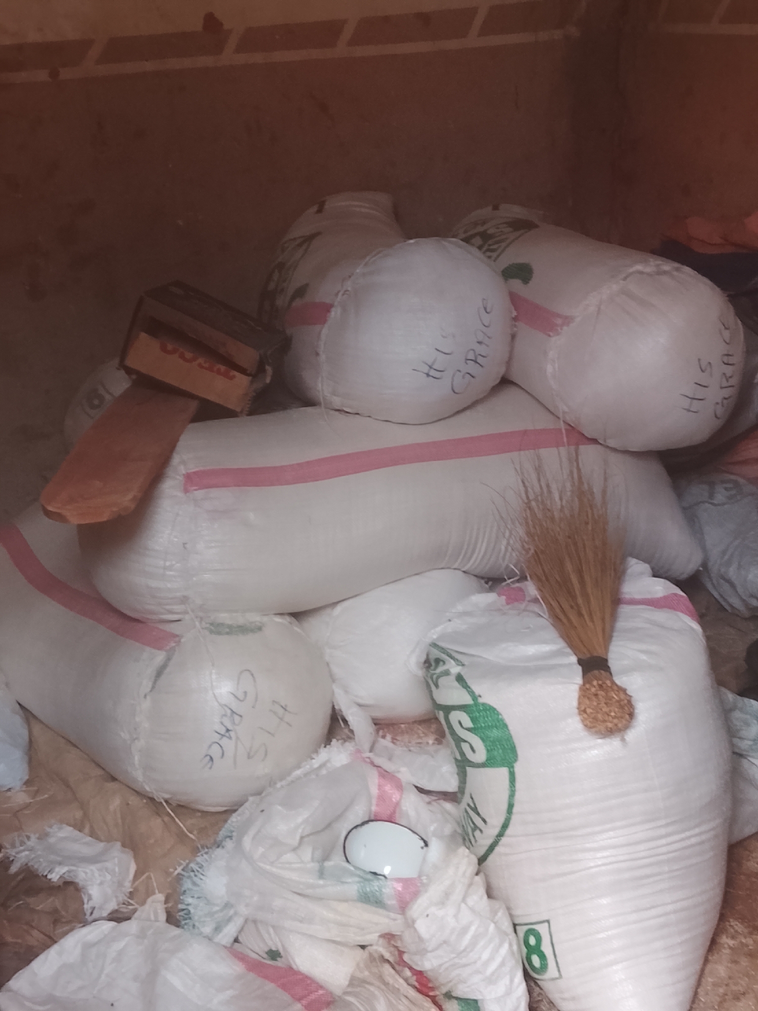 Location: Gwong, Nigeria.
Scene: A confined storage area. Multiple white sacks are piled upon each other. Some sacks have "HIS GRACE" written on them. A broom leans against one sack. A wooden object rests on top of the sacks. Debris litters the floor.