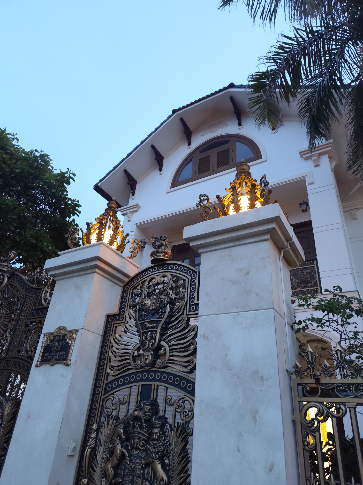 The image captures a luxurious, two-story house in Da Lat, Vietnam, characterized by its ornate, gilded gate and architectural details. The house is white with a brown, arched window on the upper floor, and dark roof tiles. Decorative lighting fixtures, shaped like crowns and emitting a warm glow, are mounted on the gateposts. The gate itself is intricate, featuring ornate designs and a plaque identifying the location as "BT YEN VY 03 KOH: DOI AN SON P3 TP. DA LAT." Foliage from nearby trees and a palm tree's fronds are visible at the sides, adding natural elements to the scene. The sky is a pale blue, suggesting early evening or dawn.