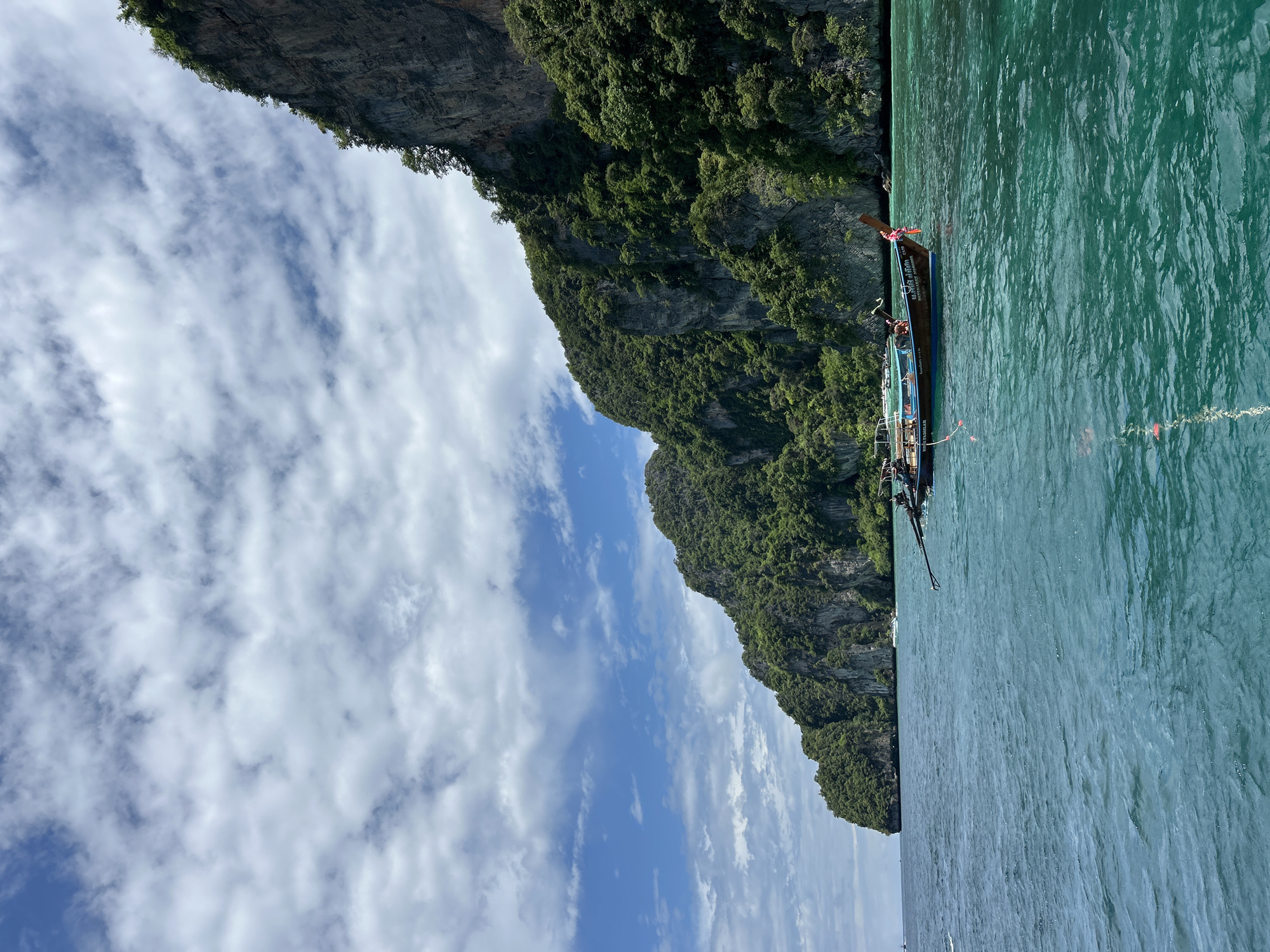This is a photograph taken from a boat on turquoise water, looking towards a densely forested, rocky island. The water is slightly choppy, with ripples and reflections. In the foreground, a longtail boat, a traditional Thai vessel, is anchored. The boat is wooden and appears to have passengers and some cargo. It is adorned with a colorful ribbon tied to its bow. In the mid-ground, other boats are visible, and further in the distance, the imposing, green-covered cliffs of the island rise steeply from the sea. The sky above is a mix of blue sky and scattered white clouds. The image conveys a sense of tropical beauty and serene maritime activity, likely in the area of Ko Phi Phi, Thailand, known for its dramatic limestone karsts and clear waters.