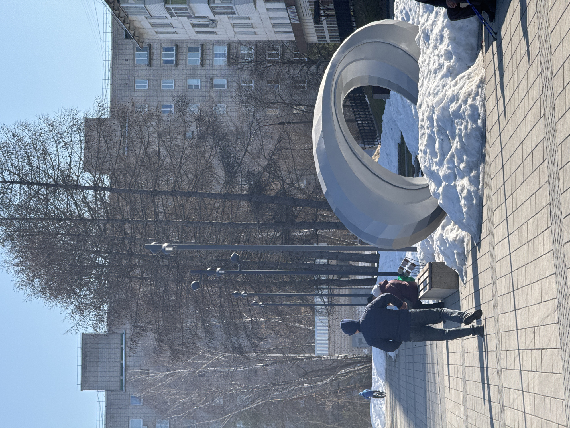 An outdoor scene shows a paved public square in Chaikovsky, Russia. A large, gray, circular sculpture dominates the right side of the frame, partially surrounded by snow. Bare trees line the background, with tall, gray apartment buildings visible behind them. A row of streetlights extends into the distance. In the foreground, a man wearing a dark jacket and jeans stands on the pavement. Behind him are two people sitting on a bench. A blue bicycle is parked at the edge of the pavement.
