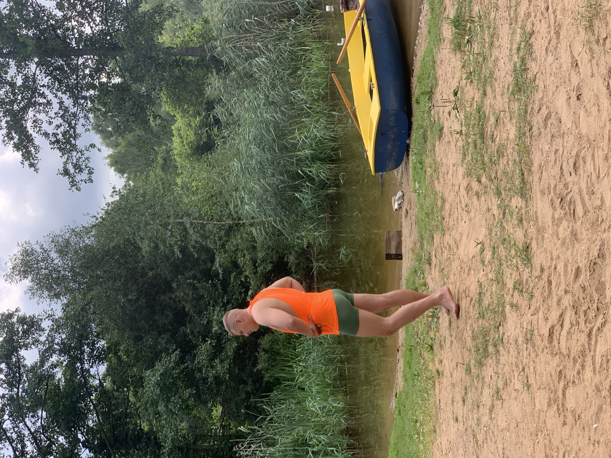A man in an orange tank top and green shorts stands on a sandy shore next to a body of water. He is facing away from the camera, with his hands on his hips. Behind him, a yellow and blue boat with oars is docked. The shoreline is lined with tall green reeds and trees. The sky is cloudy. The image appears to be taken in the Грозаўскі сельскі Савет, Belarus.