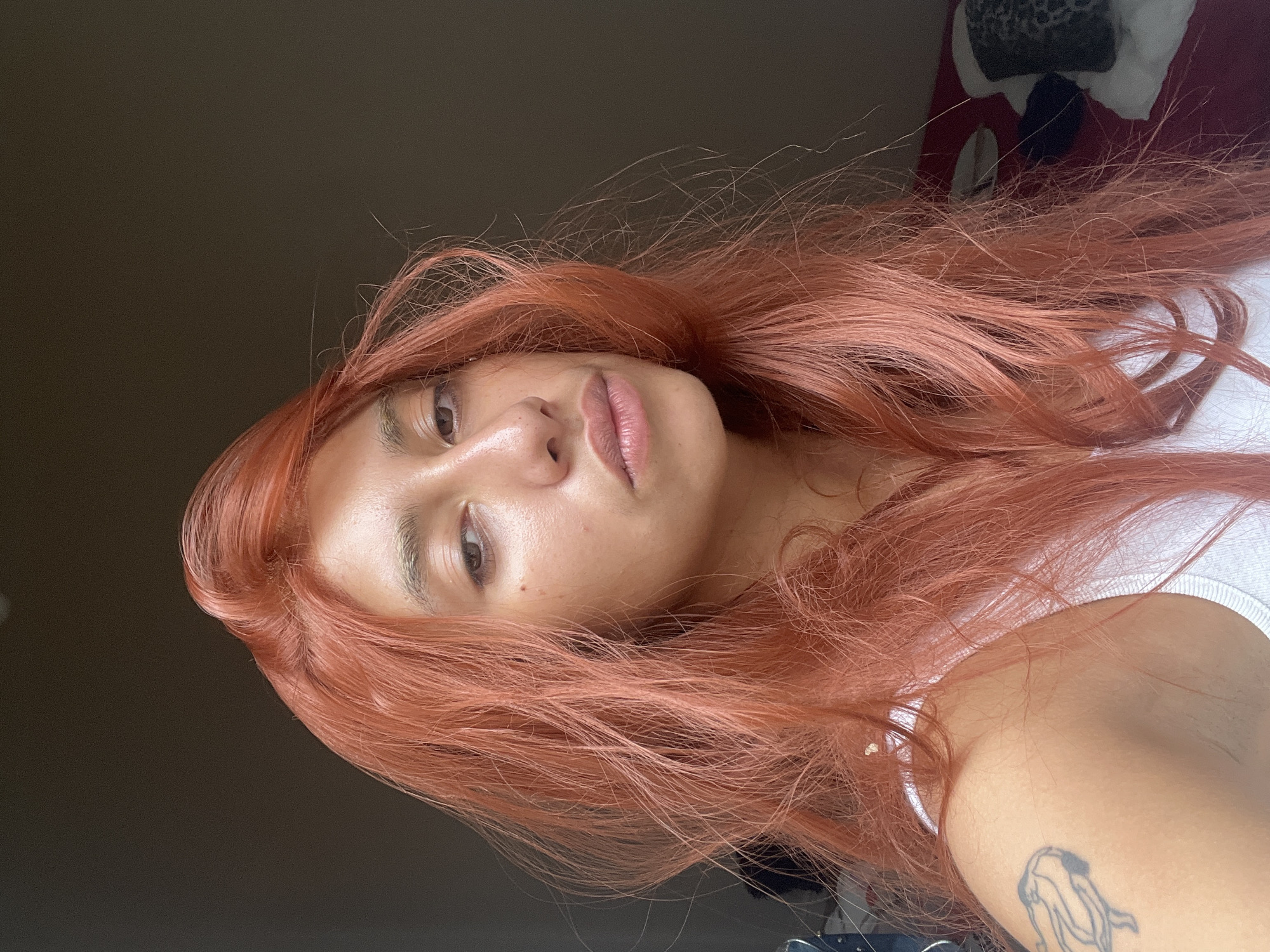 A casual, head-and-shoulder portrait, oriented vertically and rotated 90 degrees clockwise, features a young individual with striking coral-red hair. The person has light to medium brown skin and looks directly into the camera with a calm, neutral expression, their head slightly tilted. Their lips are naturally colored and softly parted. Visible features include defined eyebrows, clear skin with a slight sheen, and a small mole near the nose.
Their long, wavy hair, vibrant in its coral-red hue, frames their face and extends out of frame. They are wearing a simple white ribbed tank top. A black outline tattoo, depicting an abstract or figurative design, is visible on their upper left arm.
The background is softly blurred, indicating an indoor setting. To the left of the subject, a dark brown or grey wall is visible, while to the right, hints of white fabric, likely bedding, and a dark, patterned object (possibly a blanket or pillow) can be seen. The lighting appears soft and natural, highlighting the subject's features and hair color. The image is a self-taken photograph, suggesting a relaxed moment, and is set in Buenos Aires, Argentina. No discernible text is visible apart from the tattoo design.