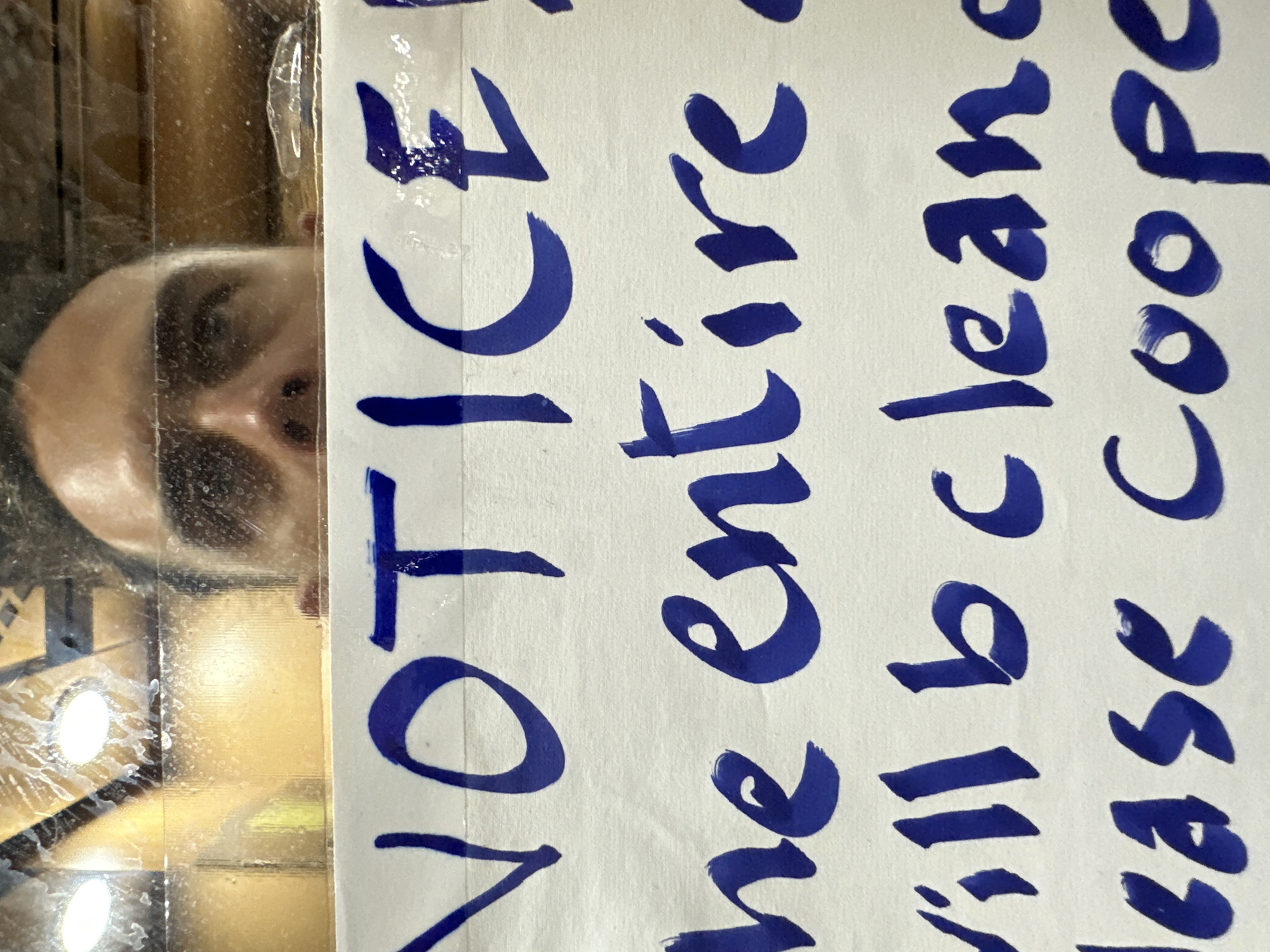 A close-up, slightly distorted, eye-level shot shows a handwritten blue sign with text reading "NOTICE! the entire will b clean. lease coop." The sign is attached to a window or glass surface, and the reflection of a person's face, specifically their eyes and forehead, is visible at the top of the image. The background behind the glass is out of focus but appears to be warm-toned, possibly indicating an indoor environment with artificial lighting. The sign itself is on white paper with visible texture. The lower part of the image shows the top of the person's head and shoulders. There are no identifiable landmarks or clues to suggest a specific location within Đà Nẵng, Vietnam.