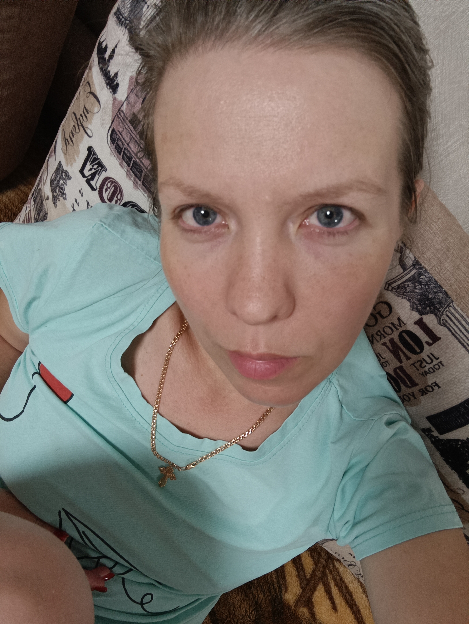 A woman is depicted in a close-up, high-angle portrait, likely taken indoors in Chaikovsky, Russia. She has fair skin, prominent blue eyes, and light brown or grayish hair pulled back from her face. Her expression is neutral, and she is looking directly at the camera.
She is wearing a light blue, short-sleeved t-shirt, which has some black line art or a graphic design partially visible on its left side. Around her neck, she wears a thin gold chain with a small gold cross pendant.
The woman appears to be reclining against a patterned pillow or cushion, which fills much of the background. This pillow features various decorative elements and text. Legible words and phrases, some appearing mirrored, include "MORNING," "LONDON," "JUST TODAY," and "FOR YOU." A silhouette resembling a double-decker bus is also part of the pattern. To her right and below her, another textured, brownish-tan surface is visible, possibly a blanket or furniture. The lighting suggests an indoor setting, and no specific time of day is discernible.