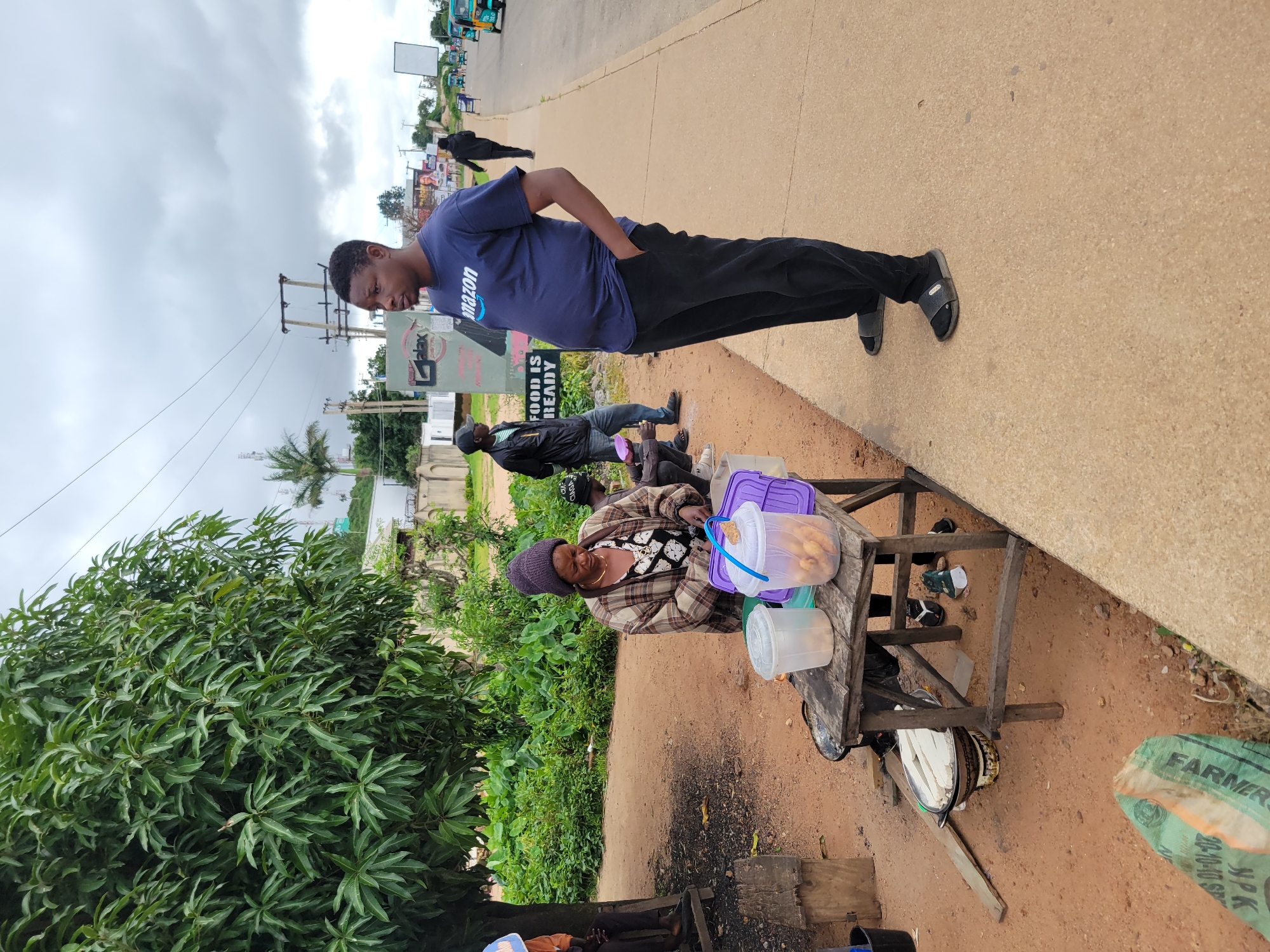 In Jos, Nigeria, on a cloudy day, a man wearing a dark blue t-shirt with the Amazon logo and black pants stands on a concrete sidewalk. To his left, a woman in a checkered jacket and a purple beanie is tending to a wooden table. On the table are two clear plastic containers, one filled with food items resembling fried plantains, and a purple container with a lid. Behind the table, a few people are visible, including a person wearing a black hoodie and another in a black baseball cap. In the background, there are lush green trees, power lines, and a billboard that reads "FOOD IS READY." Further down the street, several tricycles are seen moving along the road. The ground around the table is dusty and reddish-brown. A green bag with text, possibly fertilizer, is on the ground in the foreground.