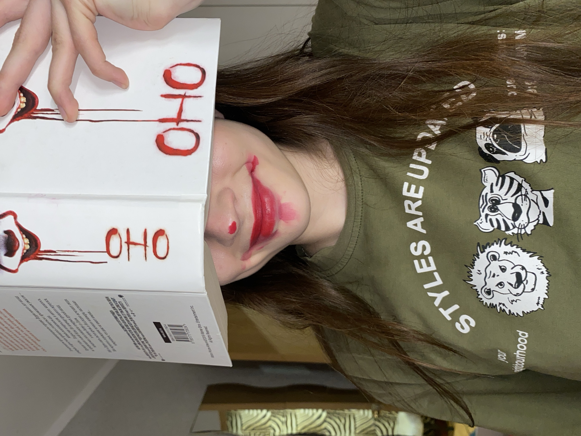 A person with long brown hair is depicted, likely lying down, with their head slightly tilted. Their face is partially obscured by a book, but their lower face is visible, adorned with theatrical red makeup resembling a joker or clown's smile, featuring smudged red lipstick and a red dot on the nose. The person has a visible smile.
They are wearing an olive green t-shirt with white text that reads "STYLES ARE UPDA" and "your neighbourhood." The shirt also features white cartoon illustrations of animal heads, including a pug, a tiger, and a lion.
The person is holding a white book over their face with their left hand. The visible part of the book cover displays disturbing red artwork, including an open mouth with sharp teeth and red lines dripping downwards, strongly resembling Pennywise from "IT." The stylized red text "OHO" is repeated prominently on the cover. The partially visible back cover contains Russian text describing a horror story, mentioning past fears and returning friends, and includes copyright information for "2019 Warner Bros. Entertainment Inc." and an ISBN (9785171175634).
The scene appears to be indoors. The overall impression is one of playful theatricality, with an unsettling, horror-themed aesthetic created by the makeup and book cover, potentially related to the "IT" franchise.