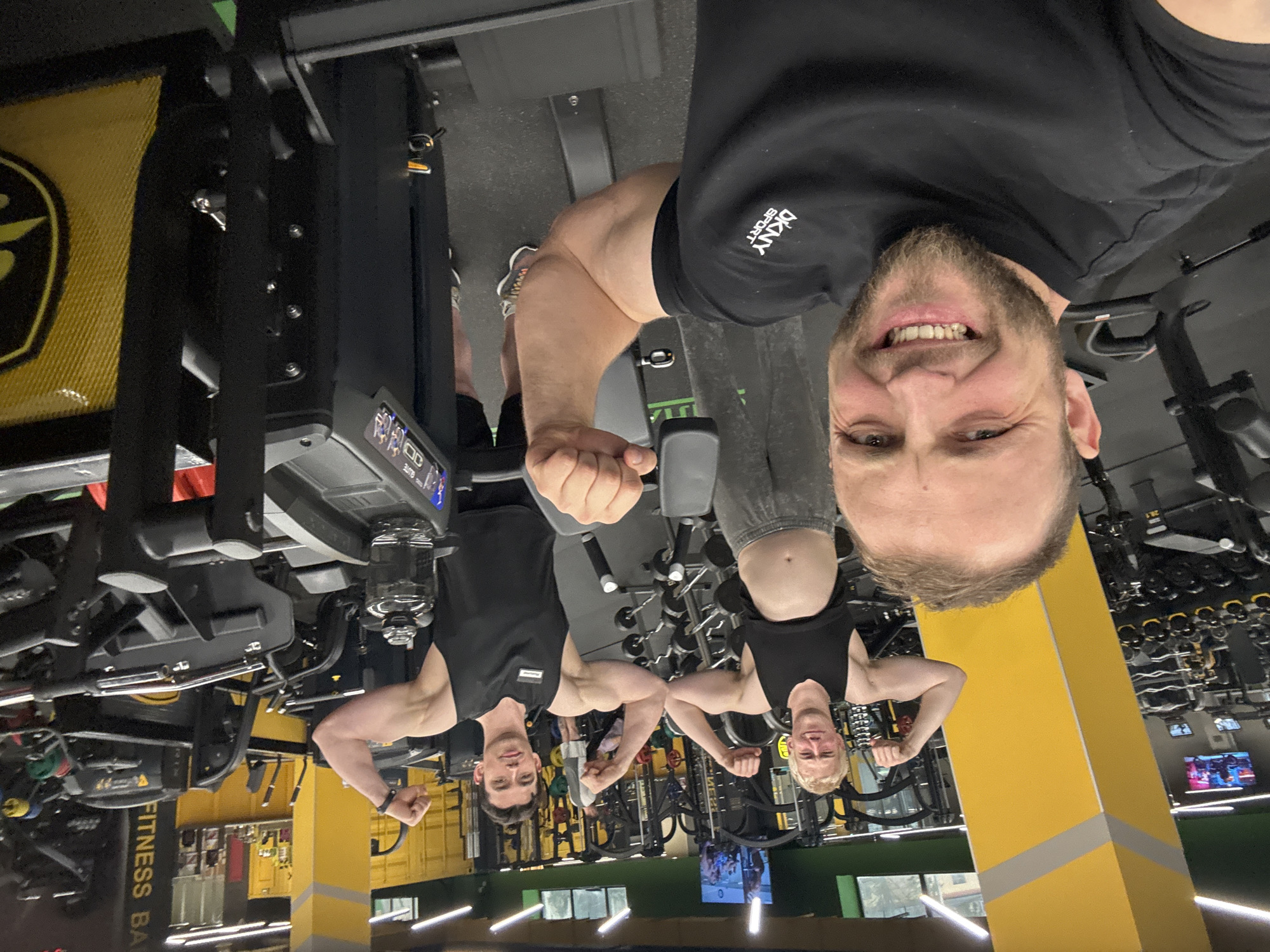 The image is a selfie taken in a gym, rotated 90 degrees counter-clockwise. The primary subject is a smiling man with a beard and short hair, looking directly at the camera. He wears a black t-shirt with "DKNY SPORT" visible on his right shoulder and is flexing his right bicep.
In the background, two other men are visible, posing and flexing their biceps. Both are wearing black tank tops; one has dark hair, and the other is blonde. They appear to be reflected, possibly in a mirror or glass surface.
The setting is a well-equipped gym with numerous machines, weight racks, and exercise equipment. The gym features a distinctive color palette of black and yellow, with yellow pillars and wall sections. On the left, a yellow banner with the partial text "FITNESS BA" is visible. The floor is dark, typical of gym flooring. Artificial lighting illuminates the entire scene, so the time of day is not discernible. One piece of equipment displays the word "ELITE" on its screen. The overall atmosphere conveys energy and satisfaction, with all subjects appearing to be enjoying their workout.