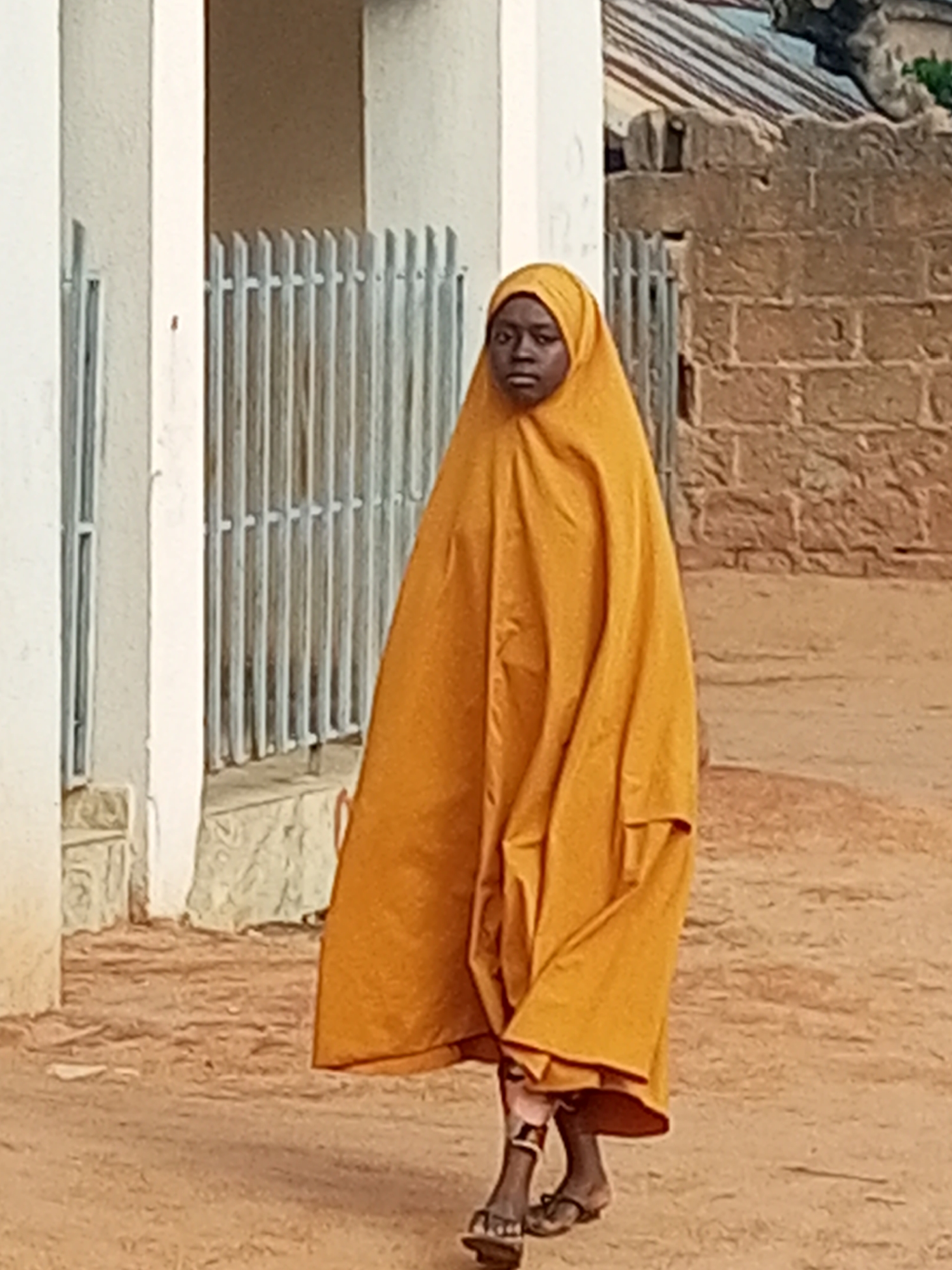 A full-shot photograph captures a young girl walking towards the camera on a dirt path in the city of Gombe, Nigeria. The girl is wearing a bright orange hijab and a matching, flowing garment that covers most of her body. Her face is visible, and she is looking directly at the camera with a neutral expression. Her feet are adorned with simple sandals.
The background features a light-colored wall with a metal railing or fence. To the right of the girl, a rough brick wall suggests a building. The ground is uneven and covered with sand and small stones. The overall lighting suggests an outdoor setting, likely during the day. The focus is on the girl, with the background slightly blurred.
