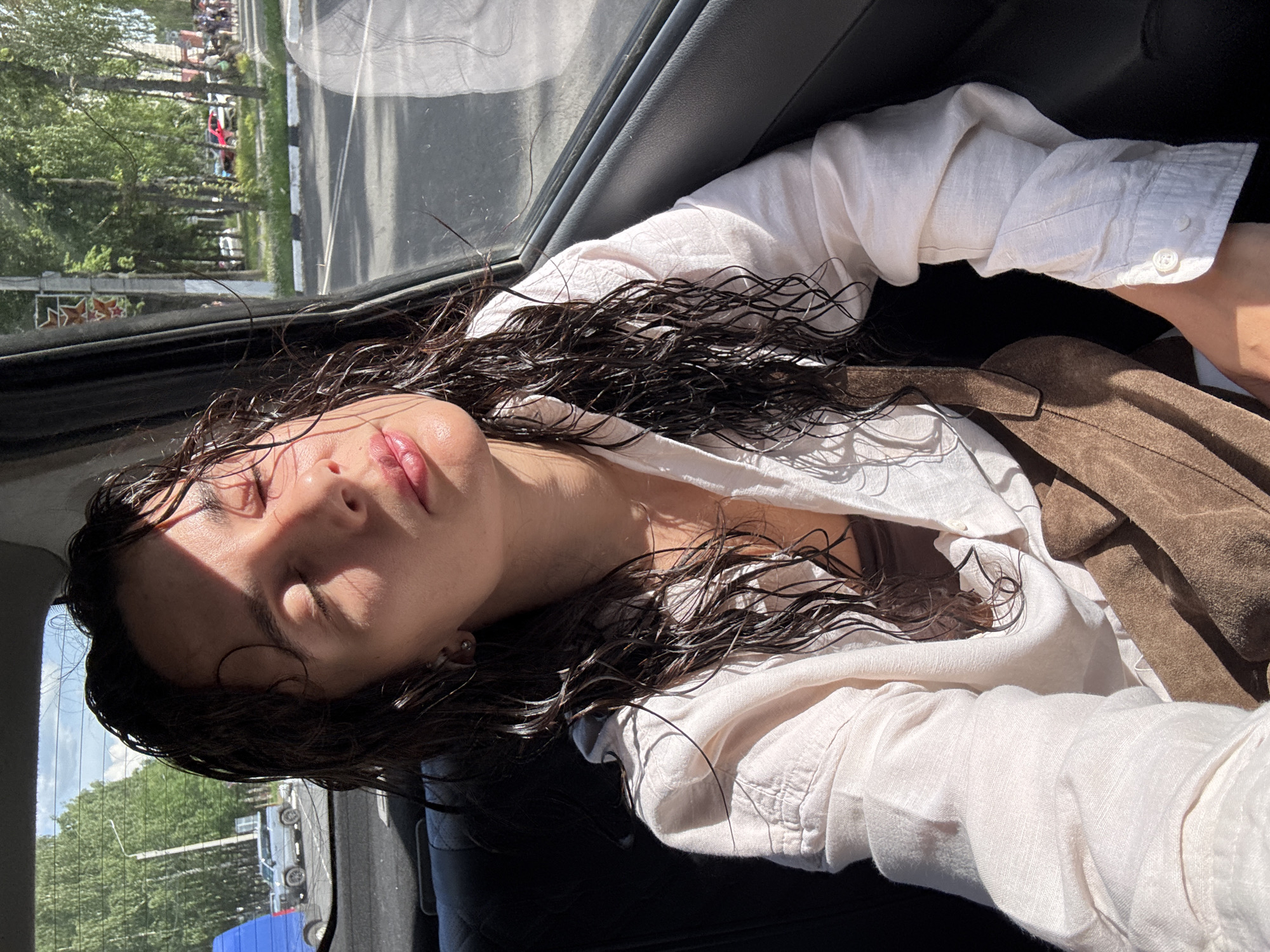 A young woman with wet, dark hair is sitting in the back seat of a car, looking towards the right side of the frame with her eyes closed. Sunlight is hitting her face, creating shadows and highlighting her features. She is wearing a light-colored, unbuttoned collared shirt over a darker top and has a brown suede bag across her chest. Through the car window, a tree-lined street in Chaikovsky, Russia is visible. The street appears to be busy with cars and pedestrians. The time of day seems to be daytime, likely late afternoon or early evening due to the low angle of the sun.