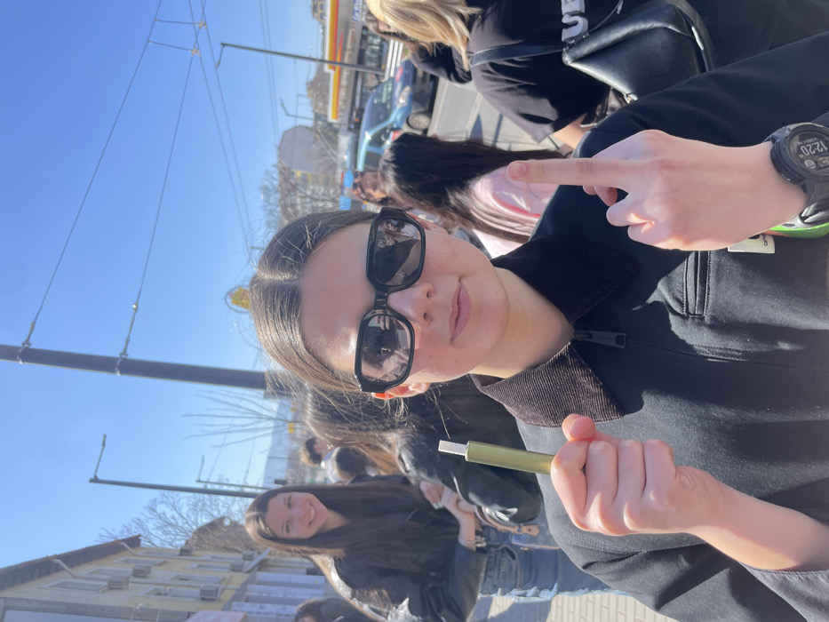 The image depicts a daytime street scene on **Bulevard "Cherni Vrah" in the Lozenets district of Sofia, Bulgaria**. The main subject is a young female centered in the foreground, looking directly at the camera. She wears dark, rectangular sunglasses and a black zip-up jacket with a dark brown corduroy collar, featuring a small white logo patch on the lower left. Her dark hair is parted in the middle.
In her right hand, she holds a slender, metallic olive-green object with a white tip, consistent with an electronic cigarette or vape pen. Her left hand is raised, displaying her middle finger towards the viewer in an offensive gesture. A black digital watch on her left wrist shows "12:20" and "21".
Behind the main subject, several individuals are present in the midground, out of focus. To the left, a young female with long dark hair, wearing a black leather jacket and blue jeans, smiles towards the camera. Other blurred figures include a person wearing a light pink top and another with a black shoulder bag.
The background reveals a clear, blue sky with bright sunlight, casting distinct shadows. To the left, a row of multi-story residential or commercial buildings lines the street, featuring windows, balconies, and visible air conditioning units. A partially visible pink sign is affixed to one building. To the right, beyond the individuals, a street with vehicular traffic is visible. Overhead wires, indicative of tram or trolleybus lines, traverse the sky above the street. In the far background, a prominent yellow Shell gas station canopy and signage are discernible, along with a light blue van on the street. Distant mountains or hills are faintly visible on the horizon. The ground beneath the subjects appears to be a paved sidewalk.