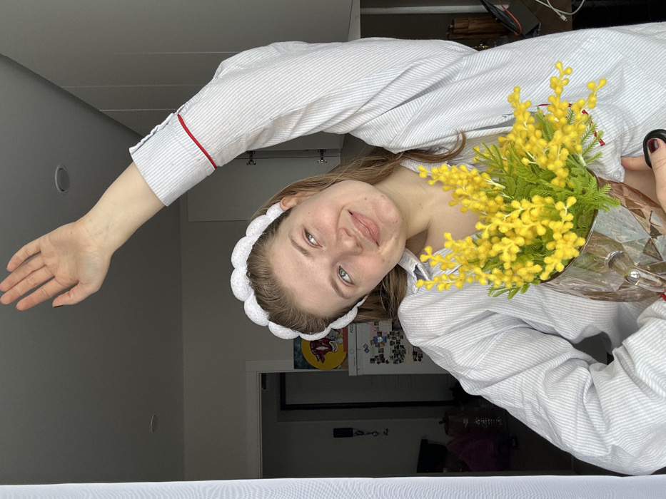 A single female subject is depicted in an indoor residential setting within Nikol'skii, Russia. The subject has light brown hair, light-colored eyes, and fair skin, exhibiting a slight upward gaze and a gentle smile. She is wearing a white, scalloped headband and a light blue and white striped long-sleeved shirt with a red trim detail on the right sleeve cuff. Her right arm is raised above her head with an open palm. In her left hand, she holds a clear, faceted glass pitcher containing a bouquet of bright yellow flowers with green foliage, consistent with mimosa. The background features a white window frame with a sheer white curtain on the left. To the right, white kitchen cabinetry with dark handles is visible. Further in the background, a refrigerator adorned with magnets and a circular colorful artwork, possibly depicting a bovine head, are discernible.