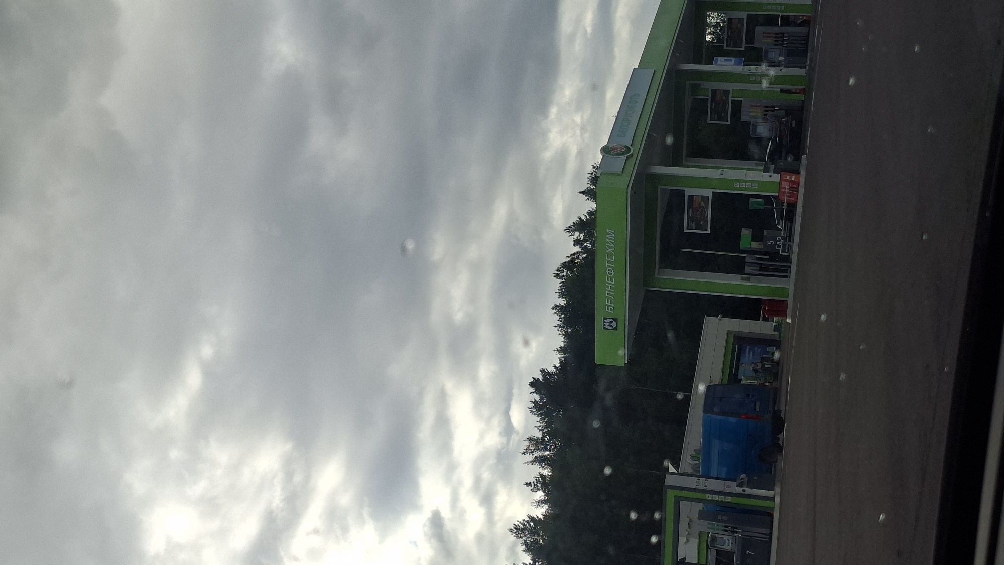 The image shows a gas station with green and white canopies. Several gas pumps are visible under the canopies. A blue van is parked to the left of the frame. In the background, a dense line of trees forms the edge of a forest. The sky is overcast with dark gray clouds. The gas station appears to be branded with "БЕЛНЕФТЕХИМ" and "БЕЛОРУСНЕФТЬ", suggesting it is part of a Belarusian oil company. The scene is likely located in the Асіпавіцкі сельскі Савет, Belarus, given the branding and typical appearance of gas stations in the region.
