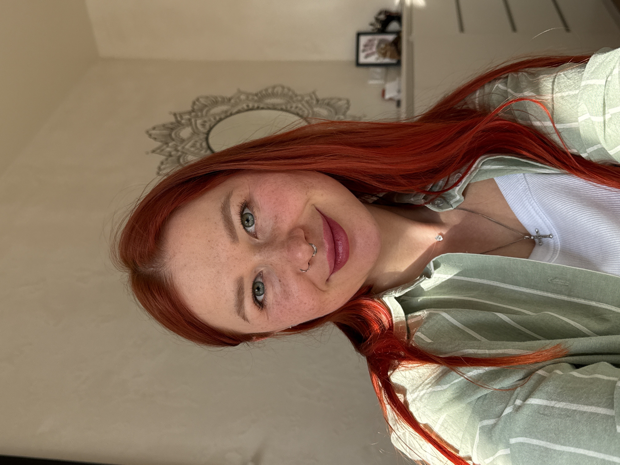 A close-up portrait features a young woman with vibrant, long red hair, fair skin, and numerous freckles, taken indoors in Rostov-on-Don, Russia. She has striking light blue-green eyes and is captured with a gentle, pleasant smile. Her lips are a soft pink. She has a septum piercing and a ring piercing on her left nostril.
The woman is wearing a light green and white striped button-up shirt, worn open over a white ribbed tank top. She accessorizes with two silver necklaces: one features a small, clear gemstone pendant, and the other a silver cross pendant.
Natural light, likely sunlight, illuminates her from the left, creating subtle highlights on her hair and face. The background consists of a light-colored wall, possibly off-white or light beige. Behind her head, a decorative, ornate mirror or wall hanging with a mandala-like design is visible. In the upper right corner of the frame, a shelf or similar surface holds a framed picture and other indistinct objects. The overall mood conveyed by her expression is cheerful and content. No visible text is present in the image.