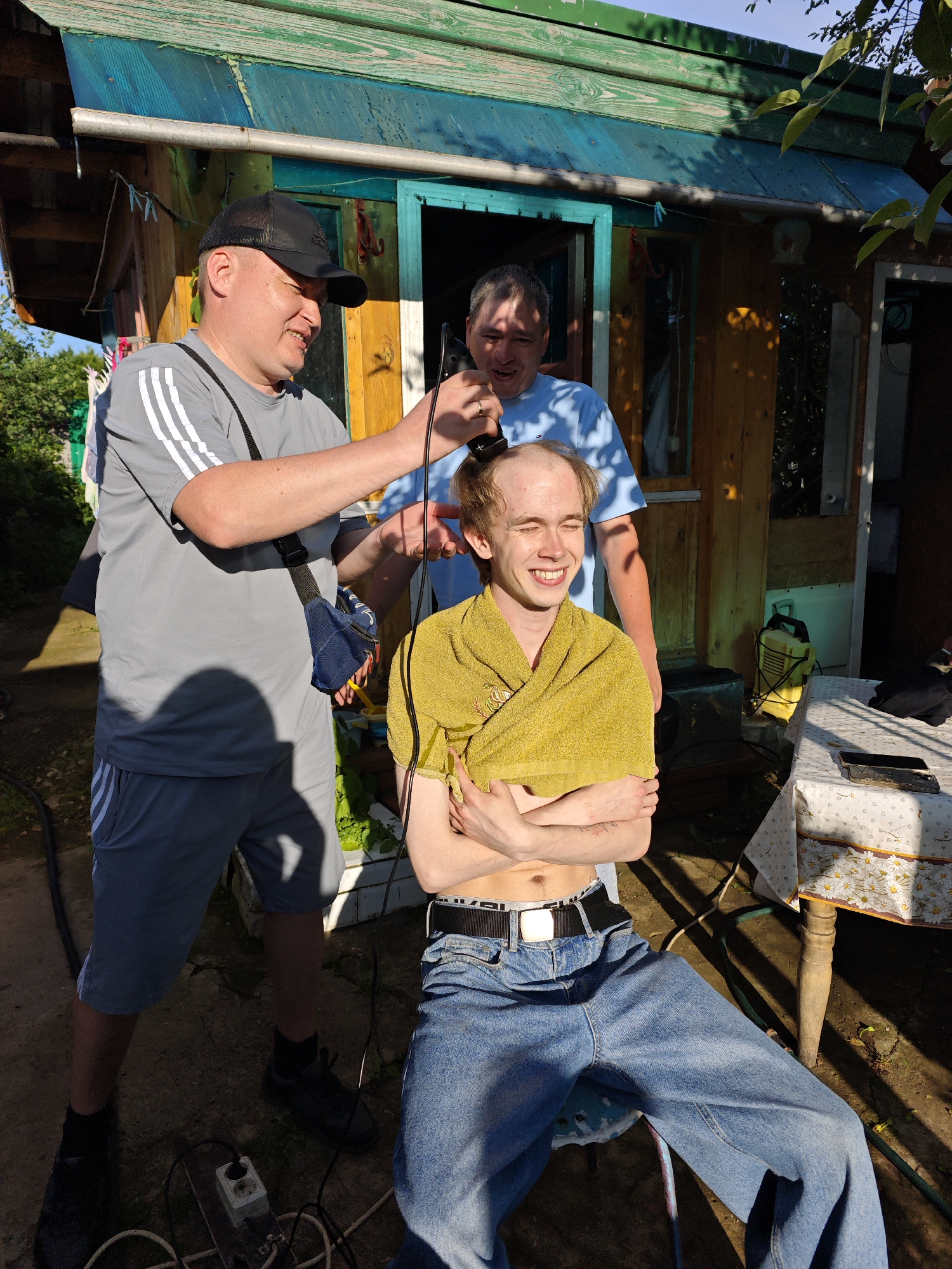 In the city of Foky, Russia, two men are on a dirt ground in front of a wooden building with a green roof and blue trim. A third man, who is bald, is being shaved by the man on the left. The man on the left is wearing a grey shirt and shorts and a black hat. The man in the middle is wearing a light blue shirt and is smiling. The bald man is wearing a yellow towel around his shoulders and is wearing jeans. The background shows plants and trees. The scene appears to be outdoors during the day.