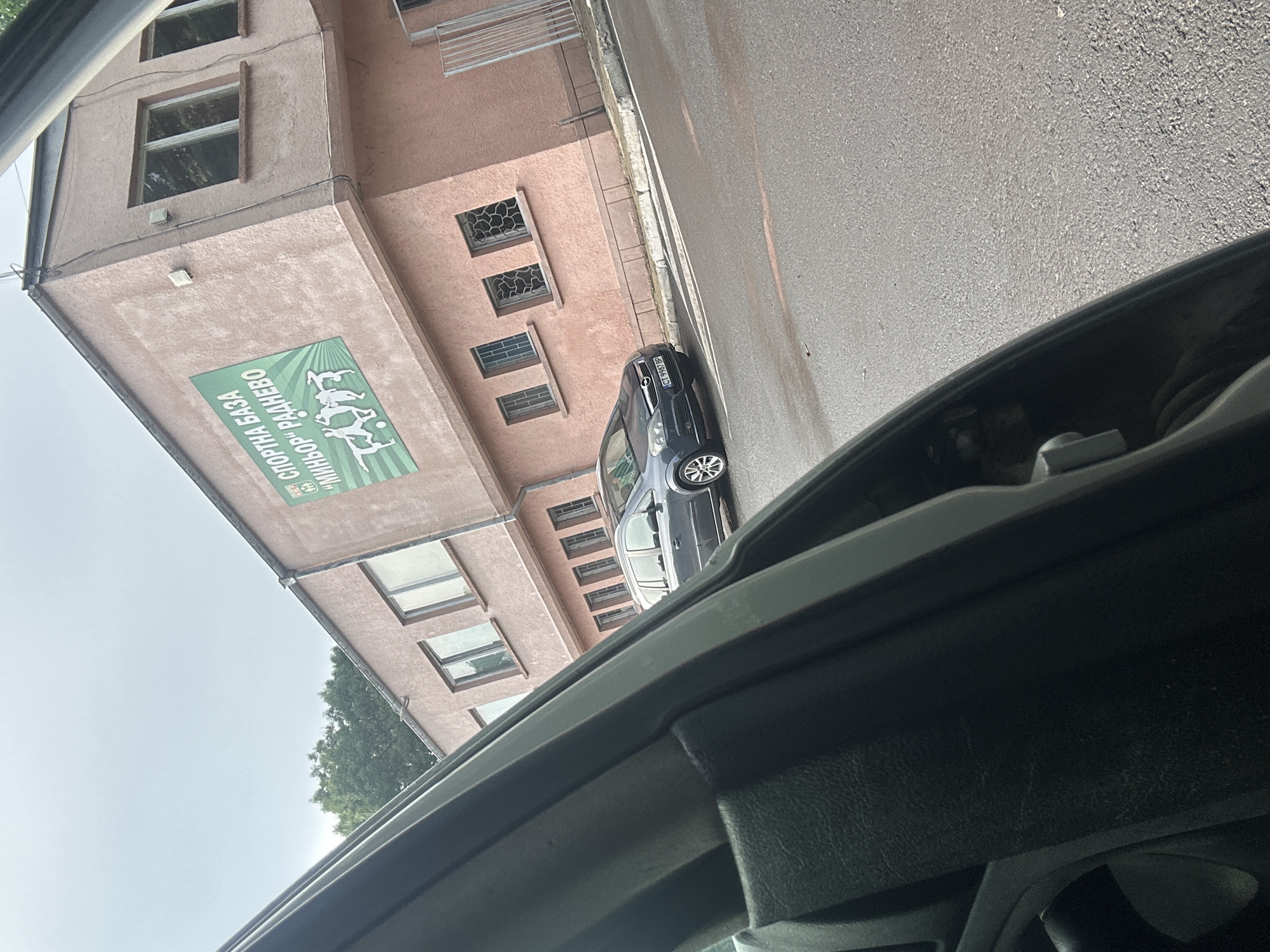 The image, taken from inside a vehicle looking out a side window, depicts a daytime street scene in Radnevo, Bulgaria. A light brown, multi-story building stands prominently on the left, featuring several rectangular windows, some with decorative metal grilles. Adorning the building's facade is a large green sign with white text, reading "СПОРТНА БАЗА" (Sports Base) and "МИНОР РАДНЕВО" (Minor Radnevo), alongside white silhouettes of various athletes engaged in sports.
Parked on the asphalt street to the right of the building is a dark gray or blue hatchback car, possibly an Opel, facing away from the camera. Its license plate, "СТ 9860 ВР", is clearly visible. A concrete curb separates the building from the street. The sky above is bright and clear, indicating a sunny day. Green tree foliage is visible behind the building in the distance. The foreground shows part of the vehicle's dark interior frame from which the photo was captured.