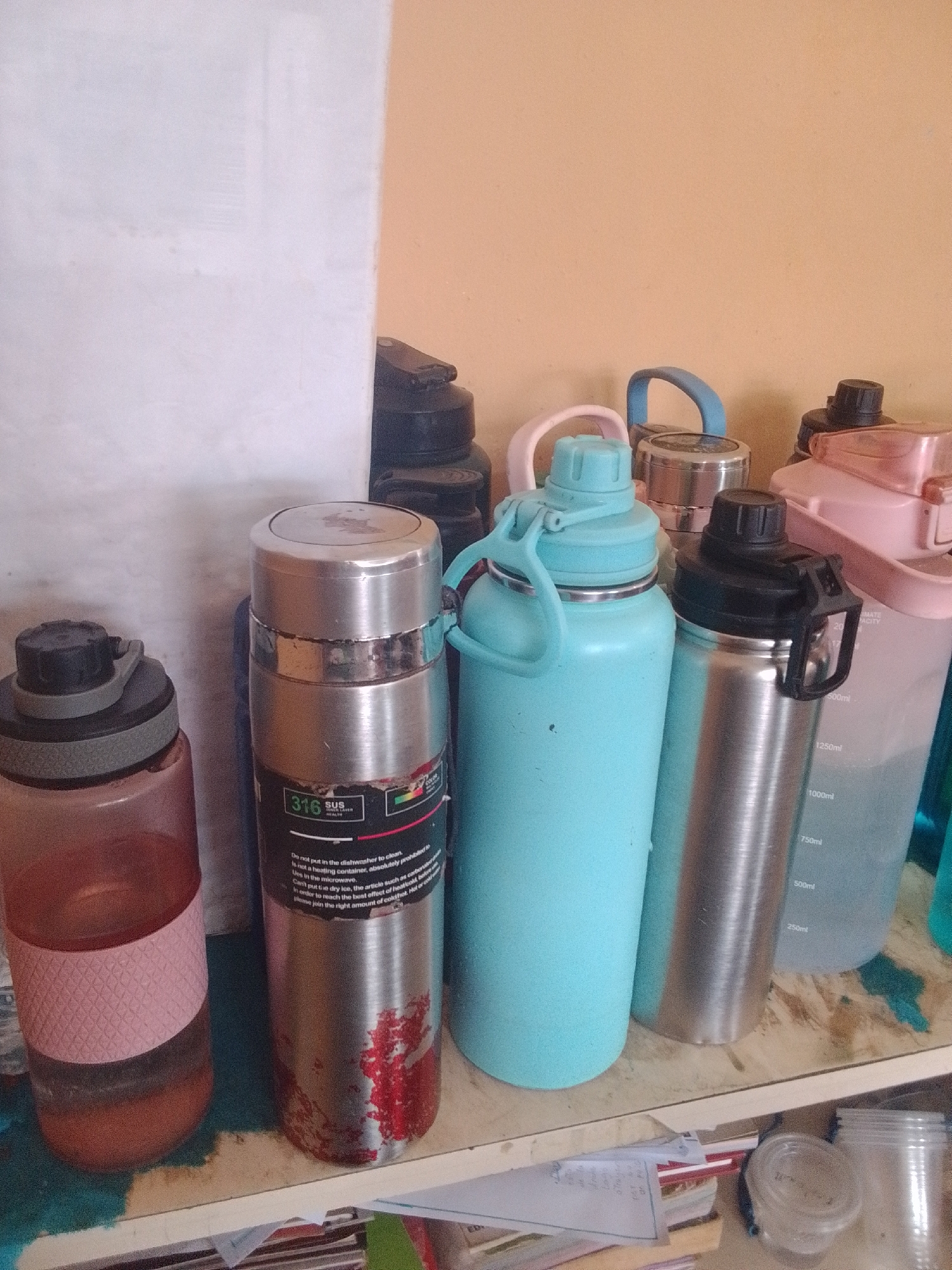 This media file depicts a collection of various reusable water bottles and containers placed on a wooden shelf. The subjects are solely objects; there are no people or animals visible. The scene is a close-up view of these items, suggesting an indoor setting, possibly a home or storage area, given the shelf and the wall in the background.
The bottles vary in size, color, and material. Prominently featured are two large, light blue and silver-colored bottles with handles and black lids. A pink bottle with a textured pattern is partially visible on the left, and a tall, silver stainless steel bottle with visible wear and red staining is in the center. To the right, a pale pink, translucent bottle with measurement markings is visible, along with other dark-colored bottles and containers further back. The lighting appears to be artificial or ambient indoor light, with no strong indications of time of day or weather.
Visible text on the silver bottle includes "316 SUS" and health-related instructions regarding cleaning and use, such as "Do not put in the dishwasher to clean. Uses in the microwave. Can't put five dry ice, the article such as carborsale In order to reach the best effect of heatcold betse Please join the right amount of cold hot Hot or colf." The pink bottle also has visible measurement markings in milliliters (ml).
The overall impression is one of organized storage of reusable beverage containers, perhaps for hydration or other purposes. The presence of wear on some bottles suggests they are actively used.