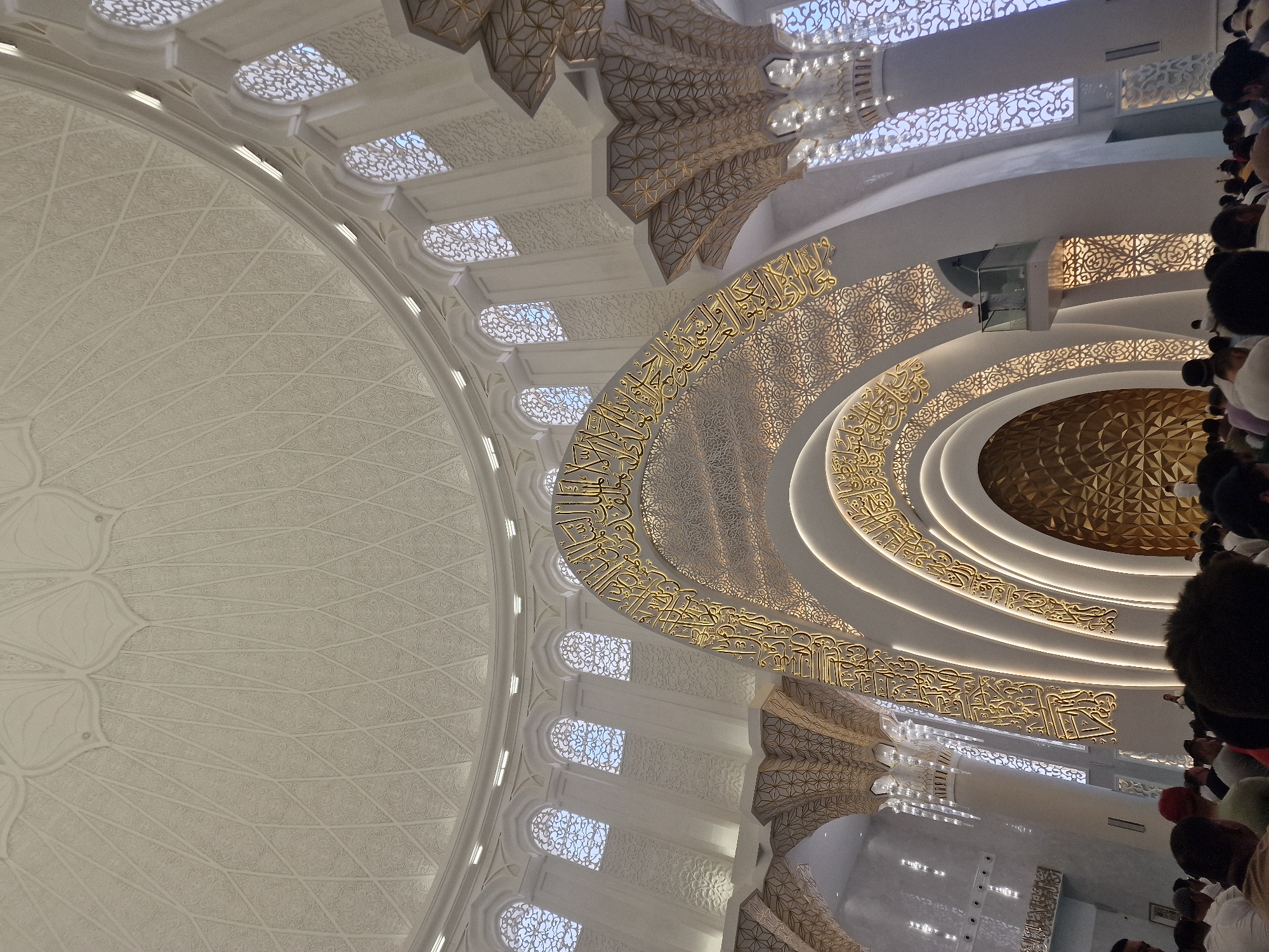 A wide-angle, low-angle shot captures the interior of the central prayer hall of the Hazrat Sultan Mosque in Shymkent, Kazakhstan. The immense, white dome, adorned with intricate geometric patterns and floral motifs, dominates the upper portion of the frame. Sunlight filters through arched, latticed windows lining the upper walls, casting a soft light on the white surfaces.
A prominent, arching structure, covered in ornate golden Arabic calligraphy and intricate geometric designs, divides the space. This arch is illuminated by warm, recessed lighting that outlines its curves. Beneath the arch, a large, golden, faceted surface forms the backdrop to what appears to be the mihrab.
Several white, fluted columns, also decorated with geometric patterns, support the arched structures and the dome. The floor is filled with a congregation of people, mostly men, dressed in white or light-colored clothing, facing towards the mihrab in prayer. The perspective emphasizes the grandeur and scale of the mosque's architecture. A small elevated platform with a railing is visible on the right side, with a lone figure standing on it, possibly an imam or speaker. The overall impression is one of serenity and spiritual reverence.