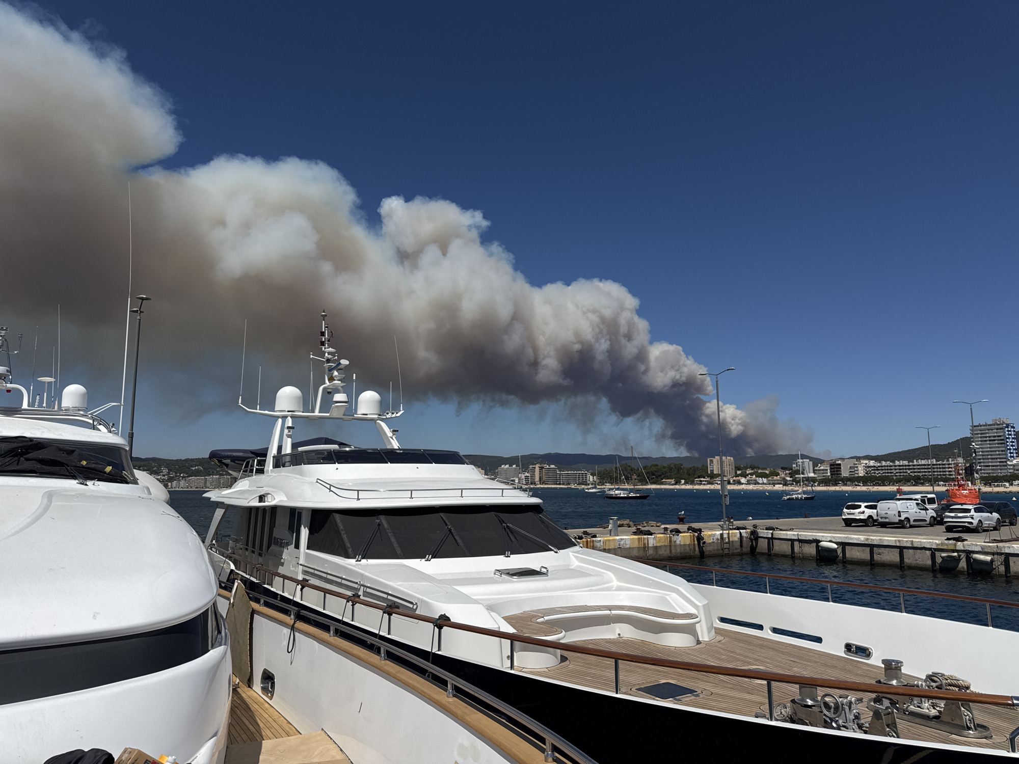 The image depicts a harbor scene in Palamós, Spain, on a clear, sunny day. In the foreground, two large white yachts are docked. The yacht on the left is only partially visible, showing its sleek white hull and a portion of its deck. The yacht on the right is more fully visible, with a polished wooden deck, white superstructure, and dark hull. Both yachts appear to be well-maintained and luxurious.
Beyond the yachts, the blue water of the harbor is visible, dotted with a few other sailboats and a small dark-hulled boat. A concrete pier runs along the right side of the image, lined with bollards and a street lamp. Several white vehicles, including cars and vans, are parked on the pier. A red and white emergency vehicle is also present.
In the background, the coastline of Palamós is visible, with buildings and trees situated on a hillside. Dominating the sky above the coastline is a massive plume of thick, dark smoke, indicating a significant fire in the distance. The smoke billows and spreads across the horizon, contrasting sharply with the clear blue sky. The scale of the smoke suggests a large wildfire or industrial fire. The overall scene captures a juxtaposition of peaceful maritime activity with the ominous presence of a distant fire.