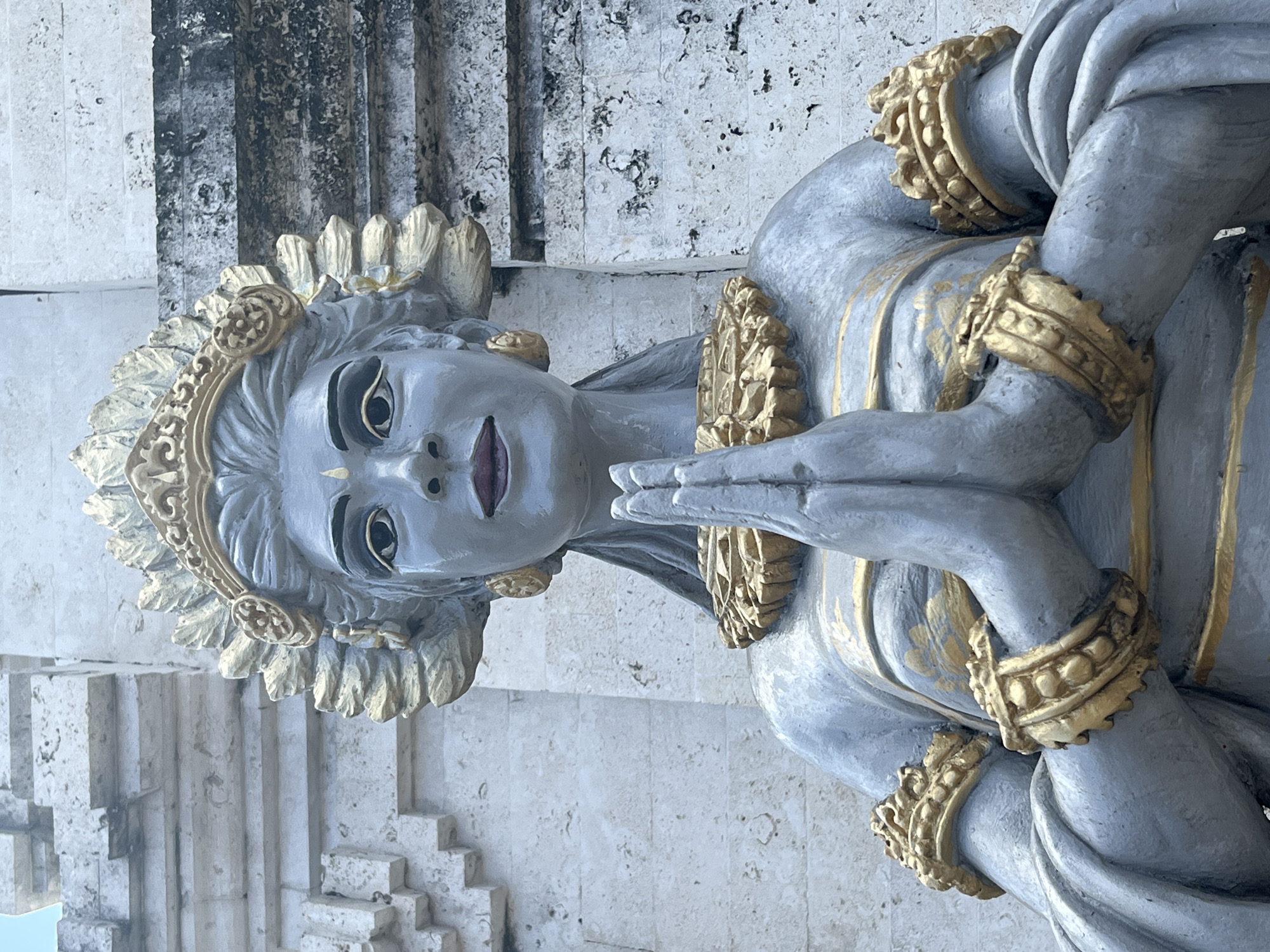 A gray stone statue of a woman with gold headdress, earrings, necklace, and armbands is positioned in front of a white stone structure, possibly a temple or building. The statue's hands are pressed together in front of her chest in a prayerful or welcoming gesture. Her eyes are wide open, and her lips are slightly parted. The background is composed of tiered, textured stone blocks, suggesting an outdoor architectural setting. The statue is a representation of Balinese culture and is located in Kuta Selatan, Indonesia, a popular tourist destination.