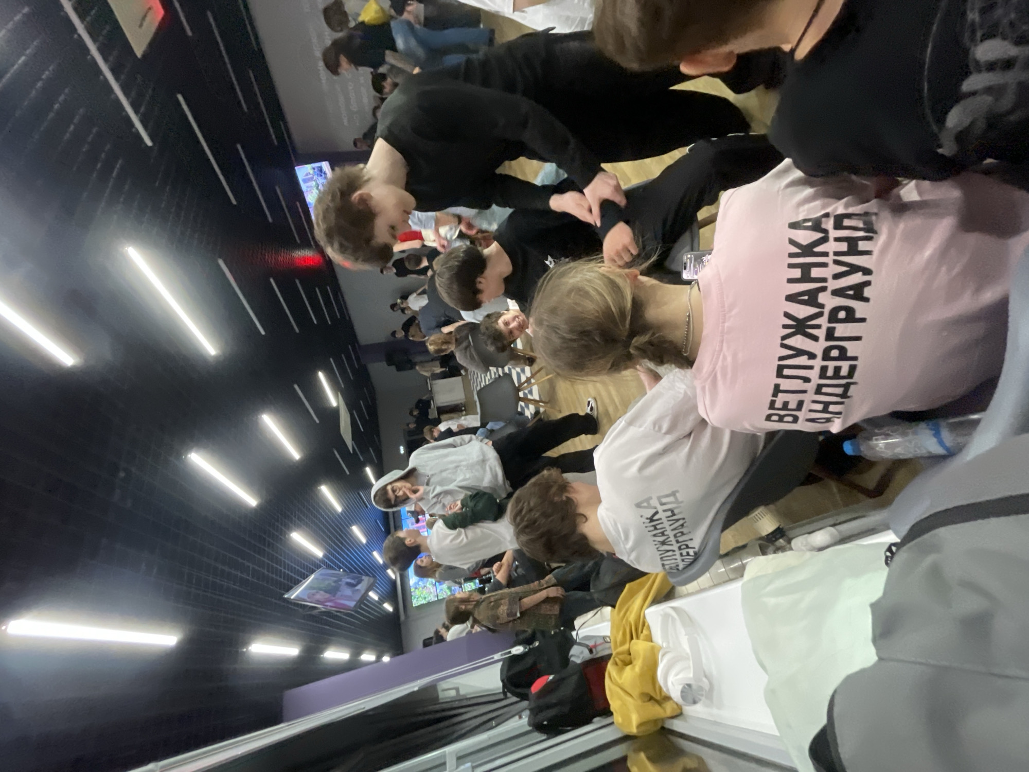 An indoor scene depicts approximately 15-20 young individuals gathered in a modern space identified as being in the Vetluzhanka district of Krasnoyarsk, Russia. This location is confirmed by the Cyrillic text "ВЕТЛУЖАНКА АНДЕРГРАУНД" (Vetluzhanka Underground) printed in black on the back of a pink t-shirt worn by a person in the immediate foreground, and on a white t-shirt worn by another individual partially visible further back.
The environment features a black ceiling with an exposed grid structure and numerous linear fluorescent or LED light fixtures. The walls are light grey, with a prominent purple accent panel on the left side of the frame. Multiple flat-screen monitors displaying digital content are mounted on the far wall. The flooring is dark.
In the mid-ground, a young male in a black long-sleeved shirt and black trousers stands, smiling, as he adjusts the left forearm of another male seated to his right, who is wearing a black t-shirt. To the left, a male in a grey hoodie and glasses looks towards the camera. Other individuals are standing, sitting on dark chairs, or interacting in small groups throughout the room.
Near a partition on the left, a black backpack, a grey backpack, and a yellow jacket are placed on the floor. White headphones rest on a white surface in the lower left corner of the image. A clear plastic water bottle is visible on the floor near the person wearing the pink t-shirt.