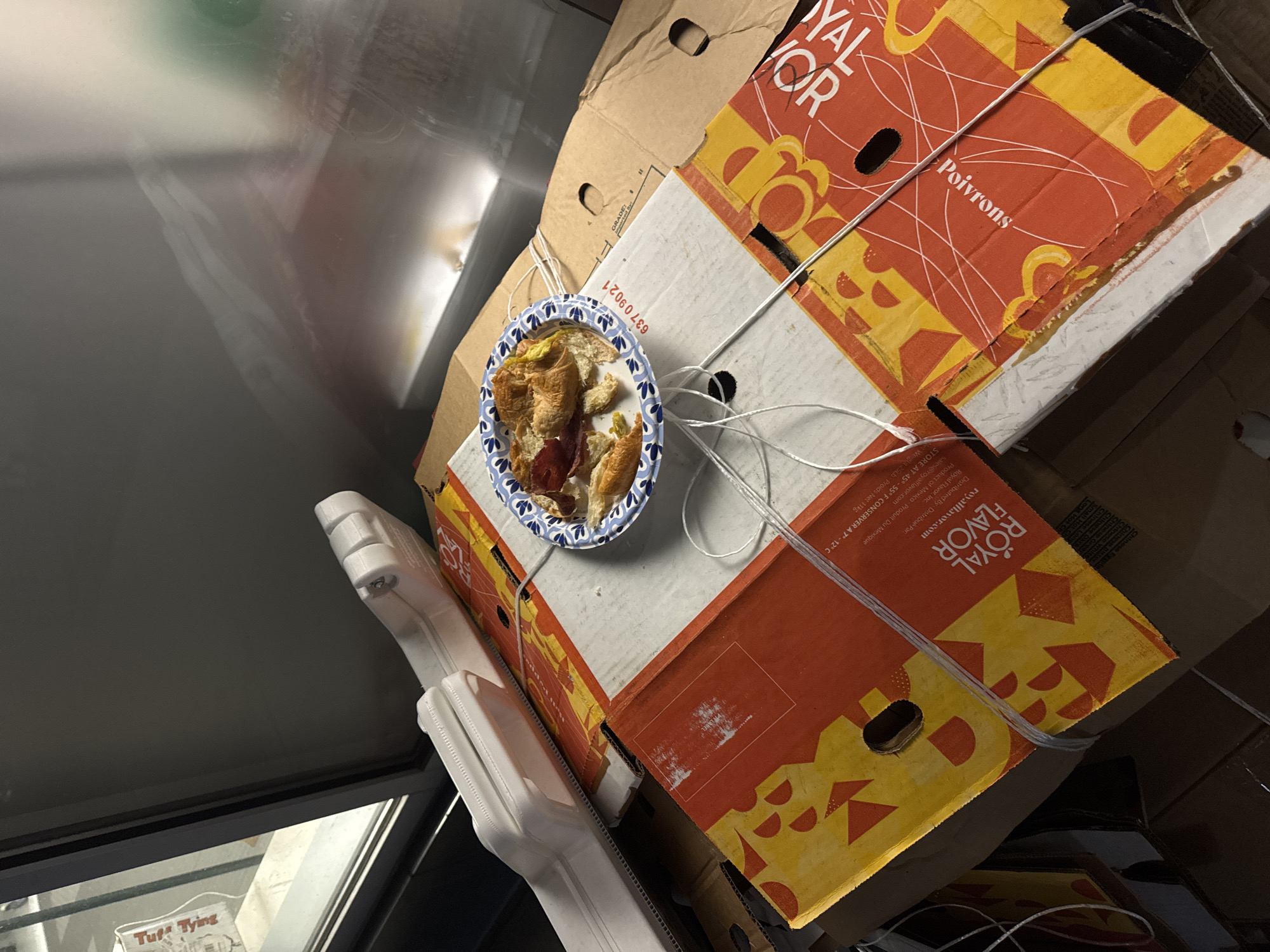 The image shows a blue and white paper plate with leftover food on top of a stack of cardboard boxes labeled "ROYAL FLAVOR Poivrons," tied together with white string. A white, unidentified plastic object is also visible. The scene is within a refrigerated unit. The reflection in the unit's door shows additional objects, and the "Tuff Trying" label can be seen in the lower left corner. The location is not evident from the image, but the "ROYAL FLAVOR" boxes and the mention of "Product of Mexico" suggests a commercial food distribution setting. The image does not provide sufficient information to identify the location as New York City.