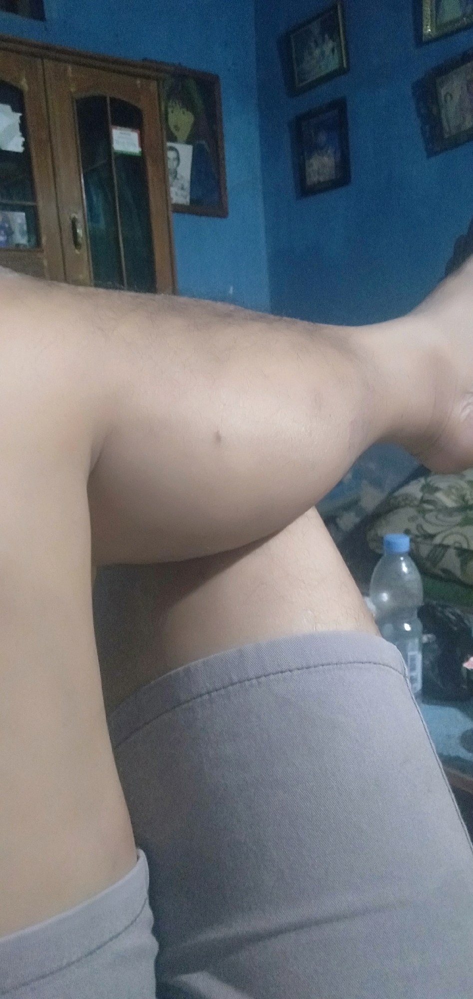 The image shows a close-up view of a person's legs, with the upper thighs and lower legs visible. The person is wearing grey shorts. The background is out of focus but appears to be an indoor setting with blue walls. There is a wooden cabinet and framed pictures on the wall. A plastic bottle of water is visible on a table in the lower right corner of the image. The lighting is dim, suggesting it might be nighttime or an indoor environment with limited light. The image is taken from a low angle, focusing on the person's lower body. The location of Plemahan, Indonesia is not explicitly visible in the image.