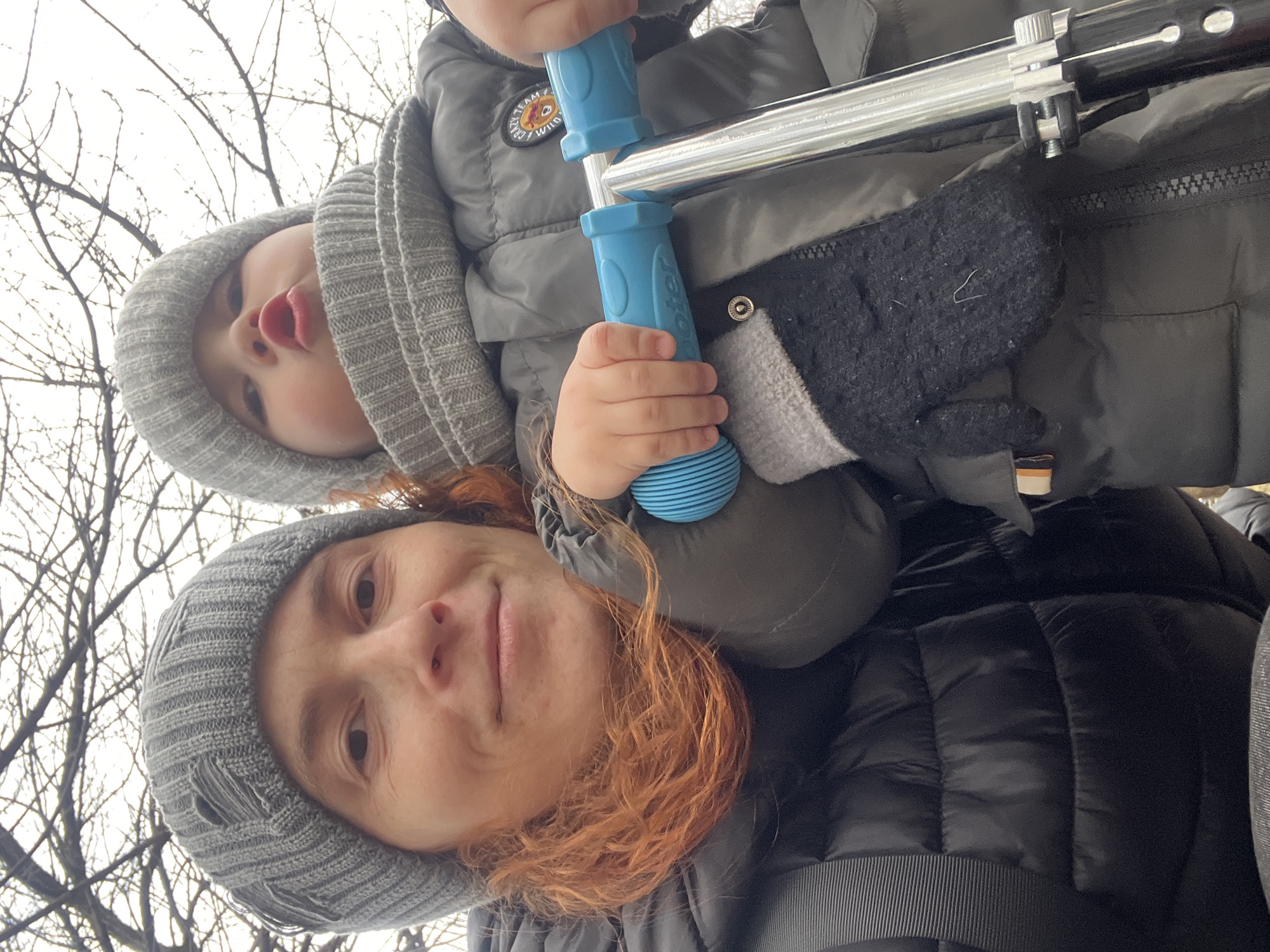 The image captures an adult and a child outdoors, viewed from a low angle. An adult, presumably a woman, with reddish-brown hair and wearing a grey knitted beanie and a dark puffer jacket, looks directly at the camera with a neutral expression. She is carrying a young child on her shoulders. The child, also wearing a matching grey knitted beanie and a grey puffer jacket, looks towards the camera with slightly puckered lips.
The child's hands are gripping bright blue handles attached to a silver metal bar, possibly part of a scooter or similar wheeled device being carried or used. The word "Scooter" is faintly visible on one of the blue grips. A patch with an unidentifiable graphic and text is visible on the child's jacket. A black mitten, possibly belonging to the adult or the child, is also visible. The background consists of numerous bare tree branches against an overcast or bright sky, indicating a colder season like winter or late autumn. The overall scene suggests a family outing or playtime in Chaikovsky, Russia.