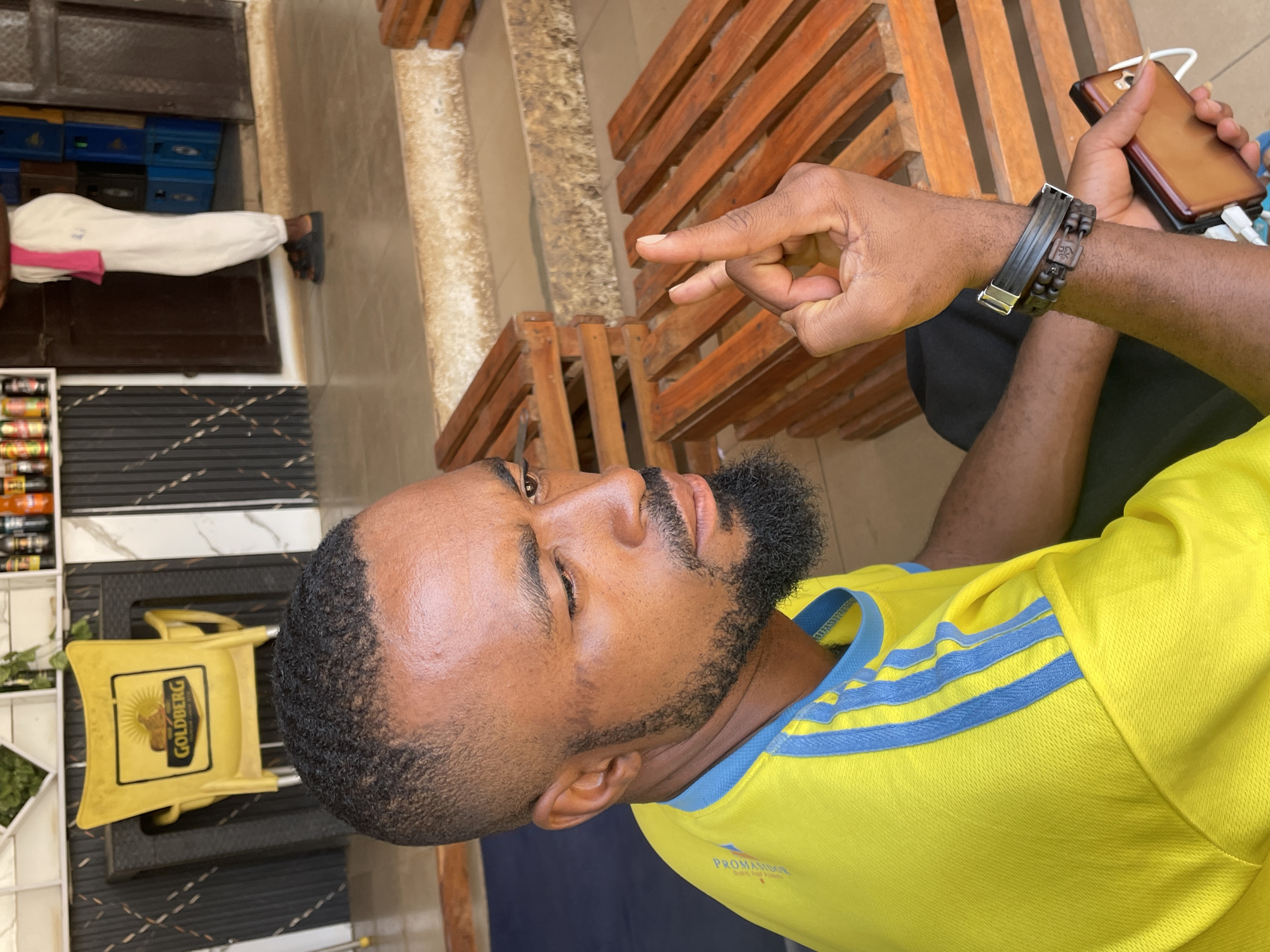 The image displays a daytime scene featuring a man with dark skin, a close-cropped hairstyle, and a well-trimmed beard and mustache. He is looking off to the upper left with a calm, pensive expression. The man is wearing a vibrant yellow t-shirt with blue stripes across the shoulders, typical of an athletic jersey, with the faint text "PROMASIDON" visible on his chest. His right wrist is adorned with two bracelets: a dark, flat band and another made of dark wooden beads with a carved charm.
His right hand is raised, with the index and middle fingers extended, while his left hand holds a brown-cased smartphone that appears to be charging via a white cable.
The setting appears to be a casual outdoor or semi-outdoor commercial establishment, possibly a local shop or bar in Barakin Akawo, Nigeria. Behind the man are wooden slatted benches, suggesting customer seating. To his left, various bottled and canned beverages are arranged on shelves. A yellow plastic container or basket, prominently displaying the word "GOLDBERG" (a Nigerian beer brand), is visible near the man. In the background, blue crates and the lower half of another person, dressed in white pants and sandals, can be seen, further hinting at a bustling local environment. The ground is tiled, interspersed with dark, textured mats. The overall atmosphere suggests a relaxed, everyday moment in a community setting.