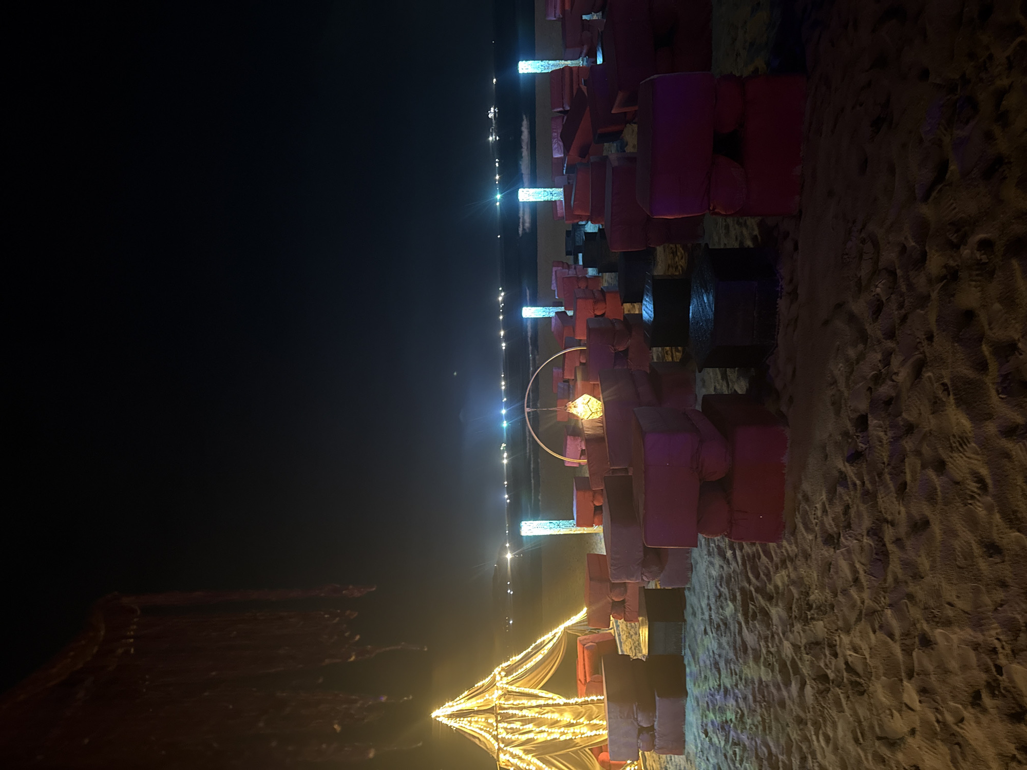 The image shows a dimly lit beach scene at night, with rows of what appear to be red and black cushioned seats or blocks stacked on top of each other along the shoreline. In the distance, there are lights scattered along the water's edge, possibly from buildings or boats. Closer to the foreground, there is a structure resembling a tent illuminated with warm, yellowish light, as well as a round, lighted hoop or decoration. The beach sand is visible in the lower portion of the image, marked with footprints. The location is Unknown, Unknown. There are no people visible in the image.