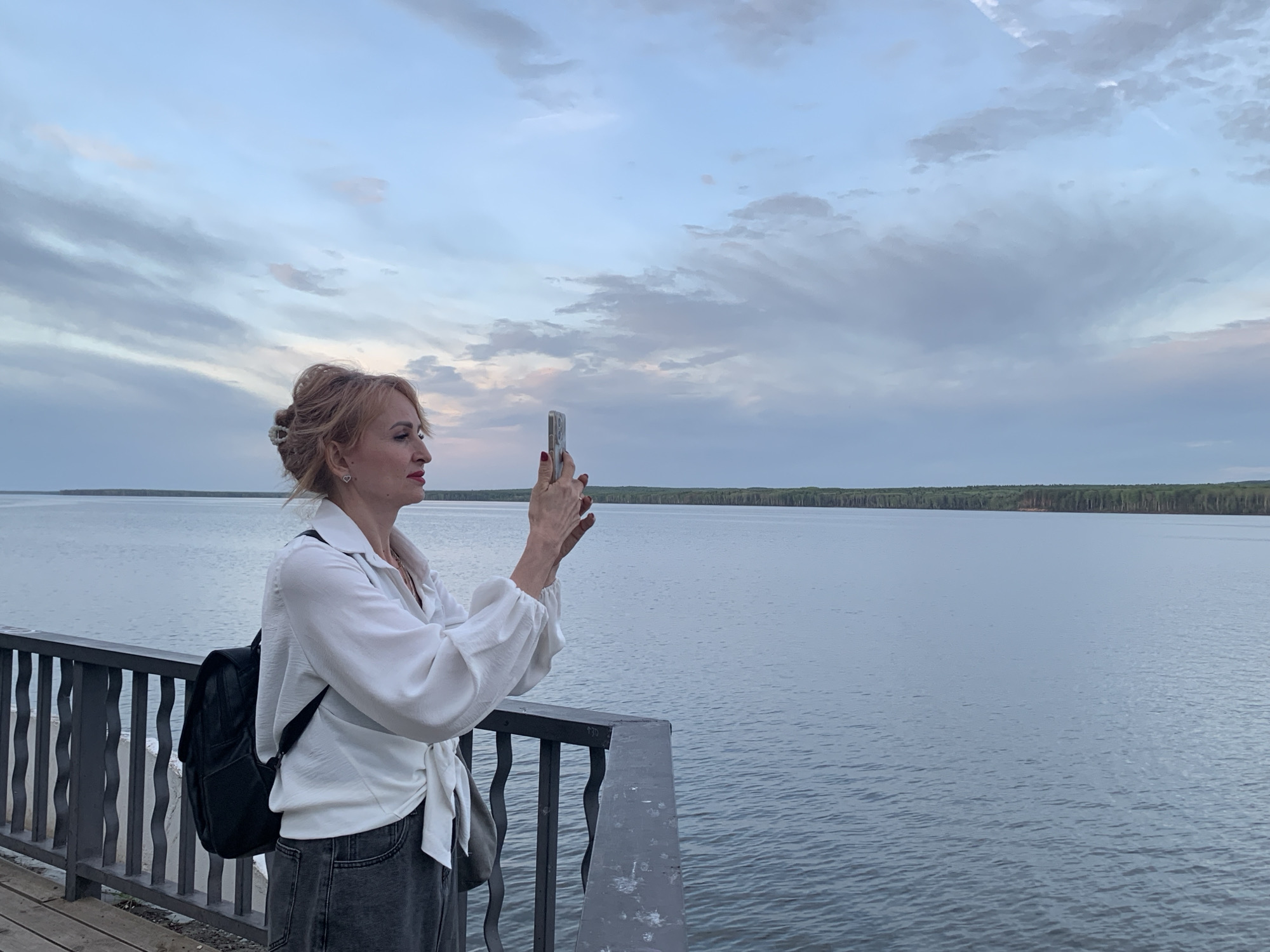 A woman stands on a pier in Chaikovsky, Russia, facing right and holding up a smartphone to take a picture of the water and horizon. She has blonde hair styled up, wears a white blouse tied at the waist, and dark jeans. A black backpack hangs over her left shoulder. The pier has a grey metal railing in the foreground. Beyond the pier, there is a large body of water with trees lining the distant shore. The sky is overcast with grey and light blue clouds.