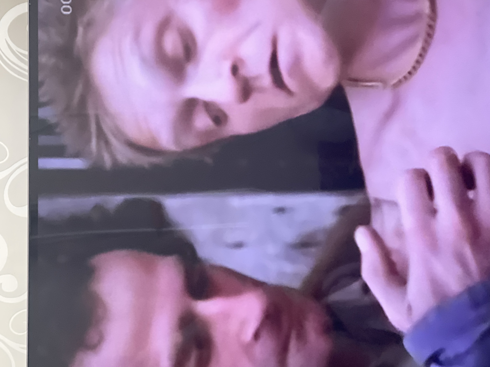 The media file is a close-up, slightly blurry image of two people. The upper subject is a person with fair hair and a fair complexion, their head tilted. They are wearing a gold-colored chain necklace. Their facial features, including eyes, nose, and mouth, are visible, and they appear to be looking downwards. A hand, likely belonging to the other person, is positioned near their face, with fingers gently touching.
The lower subject is partially visible, with their face also in close proximity to the upper subject. They have darker hair and a reddish complexion. The lighting in the image is soft, casting a pinkish hue over the subjects. The background is dark and indistinct, with some blurry shapes suggesting a surface or fabric.
There is a faint timestamp visible in the upper left corner of the image, displaying "00". No other text or discernible environmental cues are present. The overall impression is an intimate and potentially emotional moment between the two individuals.