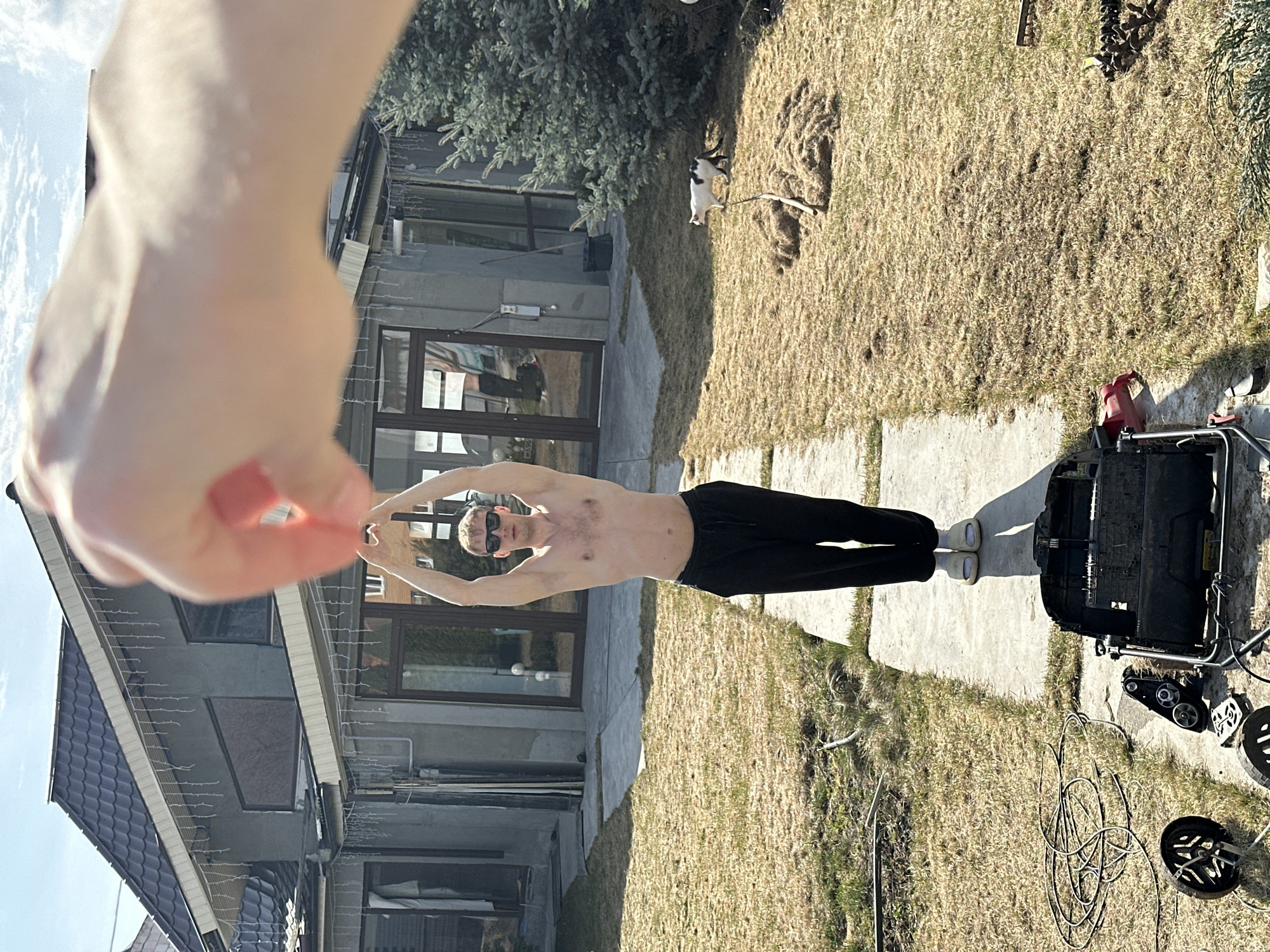 An outdoor scene shows a shirtless male with sunglasses, standing on a stone pathway in front of a house. He wears dark pants and light-colored footwear. His arms are raised overhead, and his right hand is obscured by a blurred human hand in the foreground. To his right, a disassembled piece of machinery rests on the ground alongside coiled wires and various parts. The house has a gray exterior with multiple windows and a dark roof. Grass and a bush are visible in the background, and a cat is near the grass. The image was taken outside in Kursk, Russia.