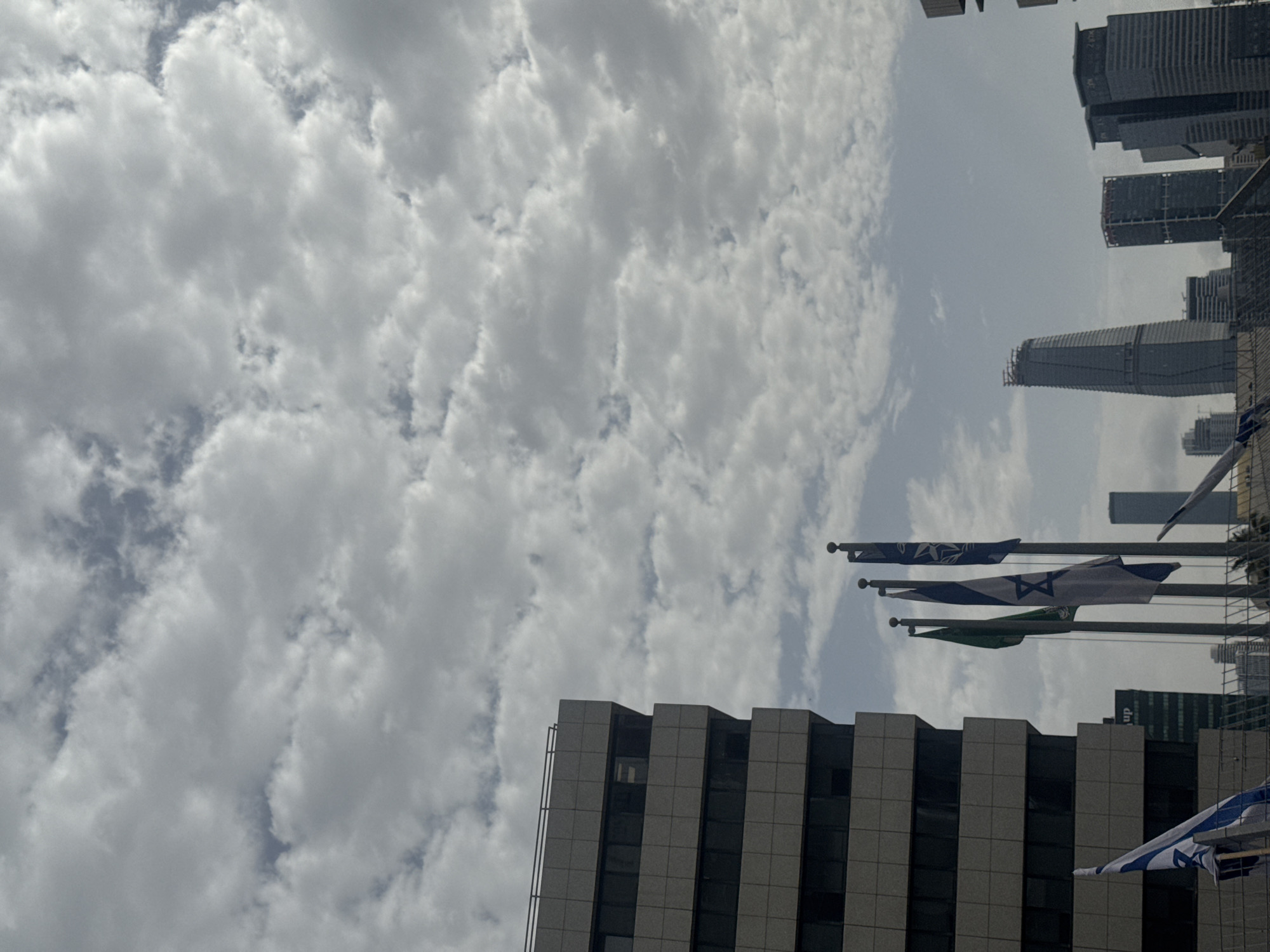 The image shows a view of the city of Tel Aviv, Israel, with a sky filled with cumulus clouds. Modern buildings and skyscrapers are visible in the skyline. Several flags of Israel hang from poles.