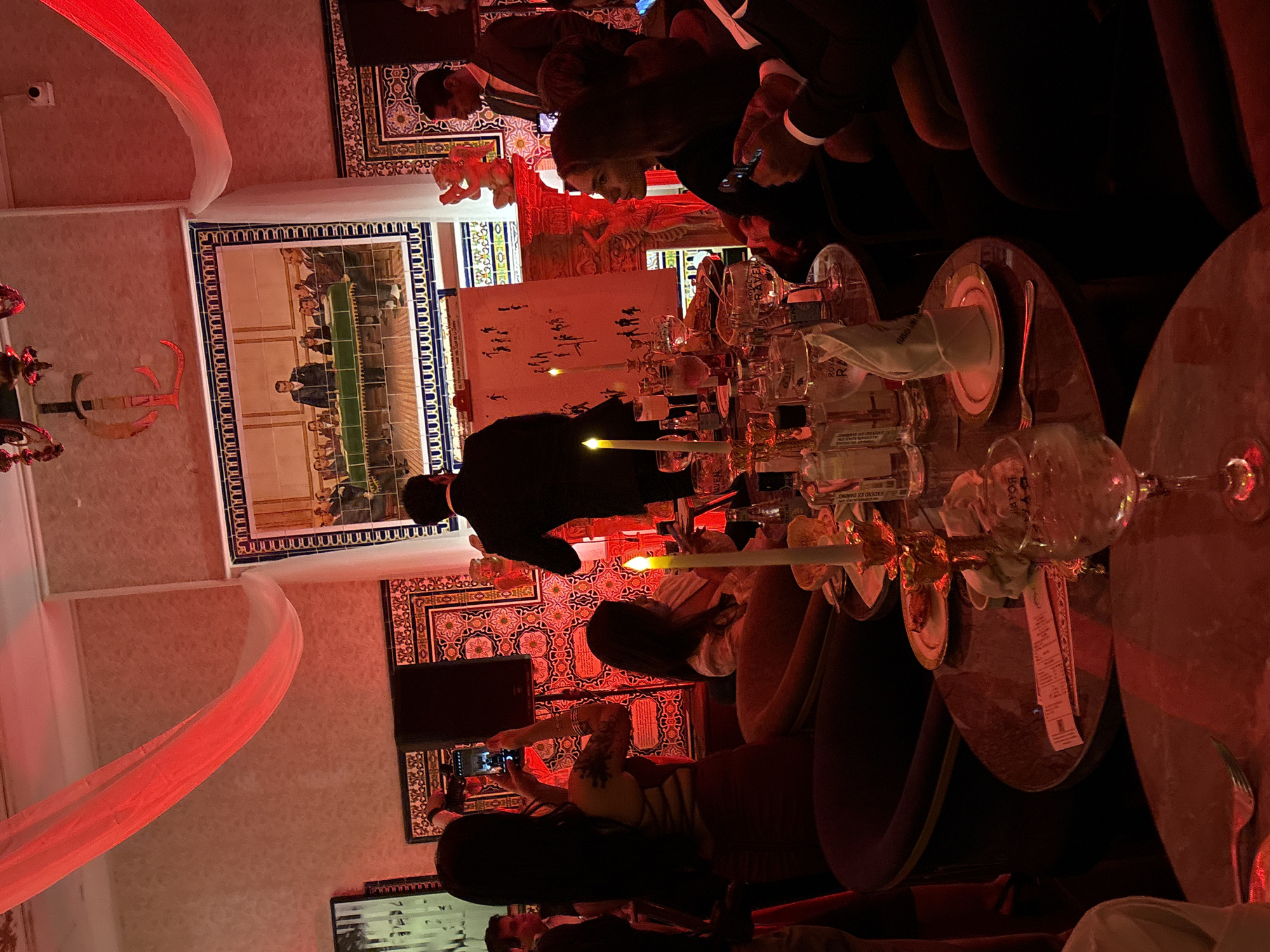 The image depicts an indoor gathering, possibly a party or event, with a distinctly red-lit ambiance. The setting appears to be a sophisticated lounge or restaurant, suggested by the presence of elegantly set tables with glassware and candles, and ornate tile work on the walls. A large framed artwork or mural hangs prominently above a decorative fireplace, depicting a group of men in formal attire around a table. Several individuals are visible, some engaged in conversation, others taking photographs with their phones, and one person is standing and gesturing towards a presentation board displaying simple stick figures. The presence of a digital camera and the way people are dressed suggests a modern event. The overall atmosphere is one of social interaction and celebration.