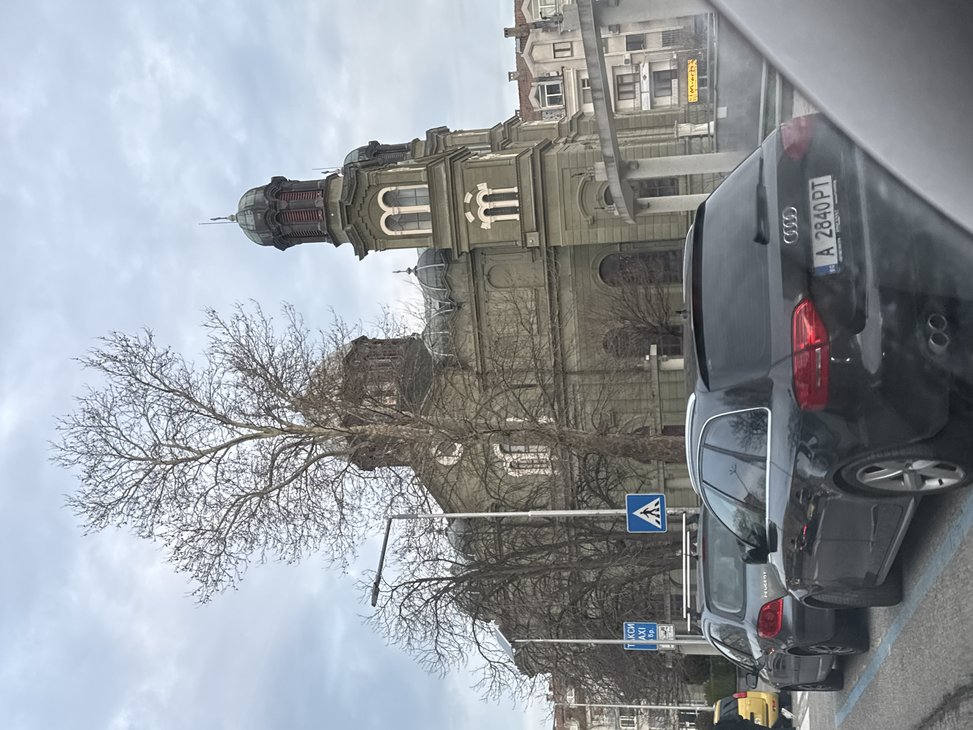 The image shows a view from inside a vehicle looking out at a street scene in Burgas, Bulgaria. The most prominent landmark is the Church of Saints Cyril and Methodius, a large green and grey domed Orthodox church. A bare tree partially obscures the view of the church. Street signs indicating a pedestrian crossing and a taxi stand are visible. Several parked cars are visible, including a dark Audi with a Bulgarian license plate reading "A 2840 PT". Other buildings are visible in the background, and the sky is overcast.