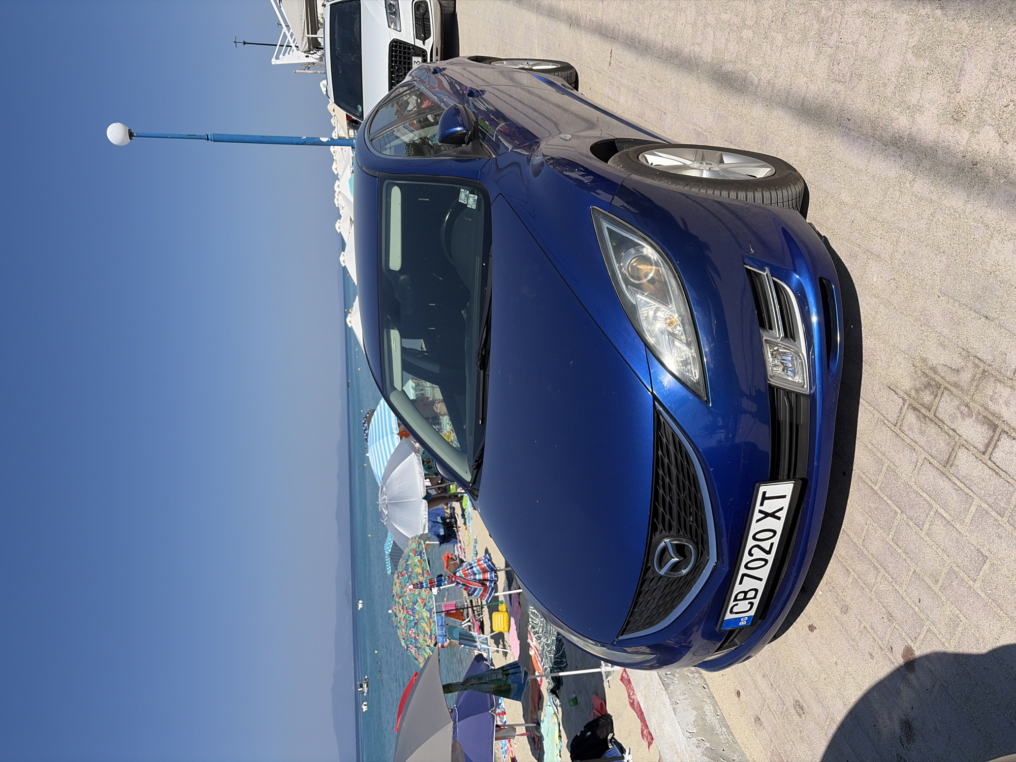 This media file shows a blue Mazda car parked on a paved area near a beach. The car is in the foreground, facing slightly to the right. Its license plate reads "BG CB 7020 XT". In the background, there is a beach with numerous umbrellas, chairs, and people enjoying the sun and sea. The ocean is visible, with calm water and distant hills. The sky is a clear, bright blue, suggesting a sunny day. A lamppost with a white globe is also visible to the left of the car. The overall scene evokes a relaxed, vacation atmosphere on a sunny day, likely in a coastal resort area. The image appears to be taken during daylight hours.
