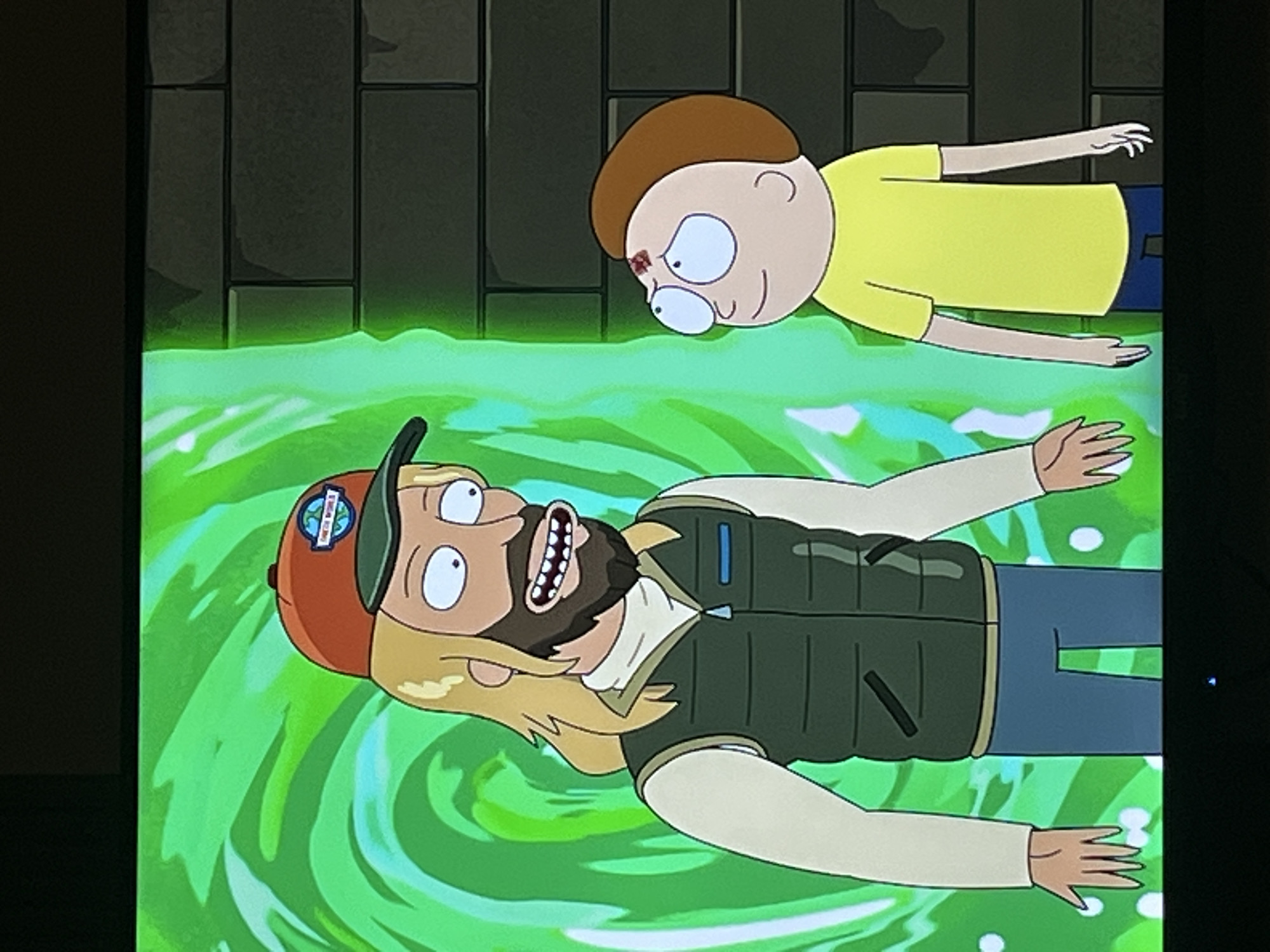 The image depicts two animated male characters in a surreal, cartoonish style, set against a backdrop of swirling green energy. In the foreground, a man with blonde hair, a beard, and an orange baseball cap with a patch faces upwards with his mouth open in a shout. He is wearing a vest over a tan shirt and blue pants. The patch on his cap appears to have text and a globe graphic. Behind him, a younger boy with brown hair and a yellow shirt looks down at him. The boy has a wound on his forehead. The green swirling background suggests a portal or interdimensional travel. The overall scene evokes a sense of urgency or danger. There are no discernible clues about the time of day or weather. The location context of Krasnoyarsk, Russia, does not appear to be visually represented in the image.