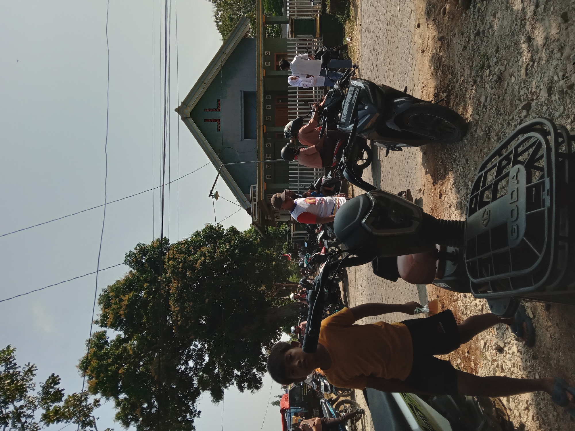 In Kedunggudel, Indonesia, on a sunny day, a young boy in a yellow t-shirt and black shorts stands on a gravelly path in front of parked motorcycles. Behind him, several other people are visible, some on motorcycles and others standing. A light green house with a pointed roof and white fence is in the background, surrounded by trees and utility wires. The scene appears to be a street or a gathering area.