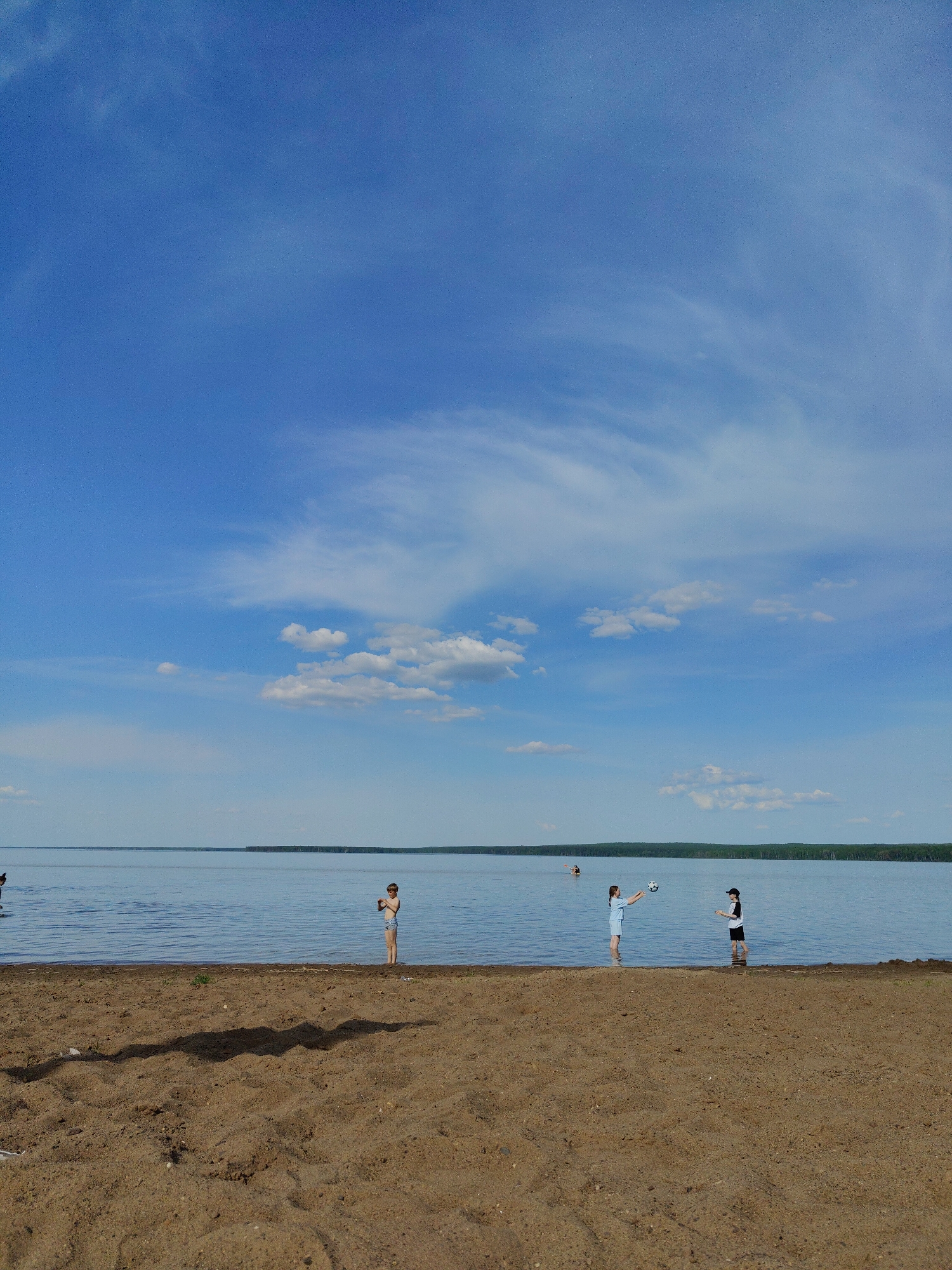 The image shows a sunny beach scene, likely on a lake or large body of water, under a bright blue sky with scattered clouds. The foreground is a sandy beach. In the water, three people are visible: one individual is standing alone further to the left, while two others stand closer to the right side, one throwing a ball to the other. A wooded area is visible in the distance across the water. The location is Unknown, Unknown.