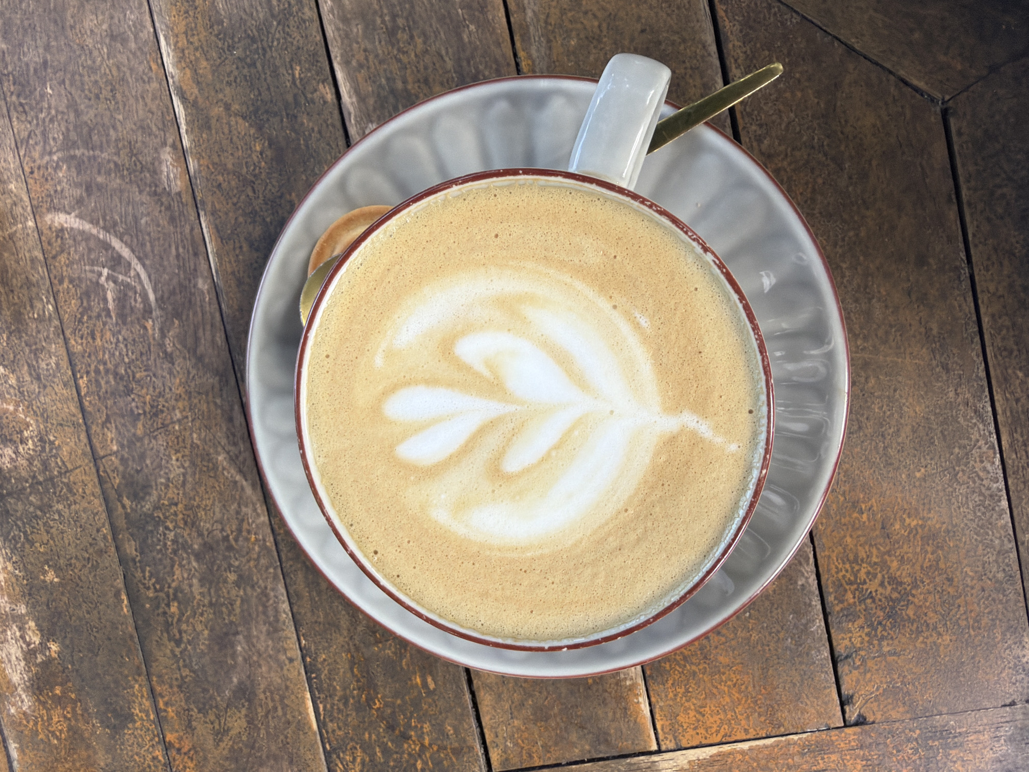 This media file is an overhead shot of a coffee cup filled with latte art, placed on a saucer. The cup and saucer are resting on a rustic wooden table. A spoon with a gold-colored handle is visible, peeking out from behind the cup. The latte art is a delicate, leaf-like pattern created with steamed milk. The wooden table has a textured, weathered appearance, suggesting it might be outdoors or in a casual setting. The lighting appears to be natural, casting soft shadows, which could indicate daytime. The scene evokes a sense of calm and enjoyment, typical of a coffee break. There is no visible text in the image. The location context is Kuta Selatan, Indonesia.